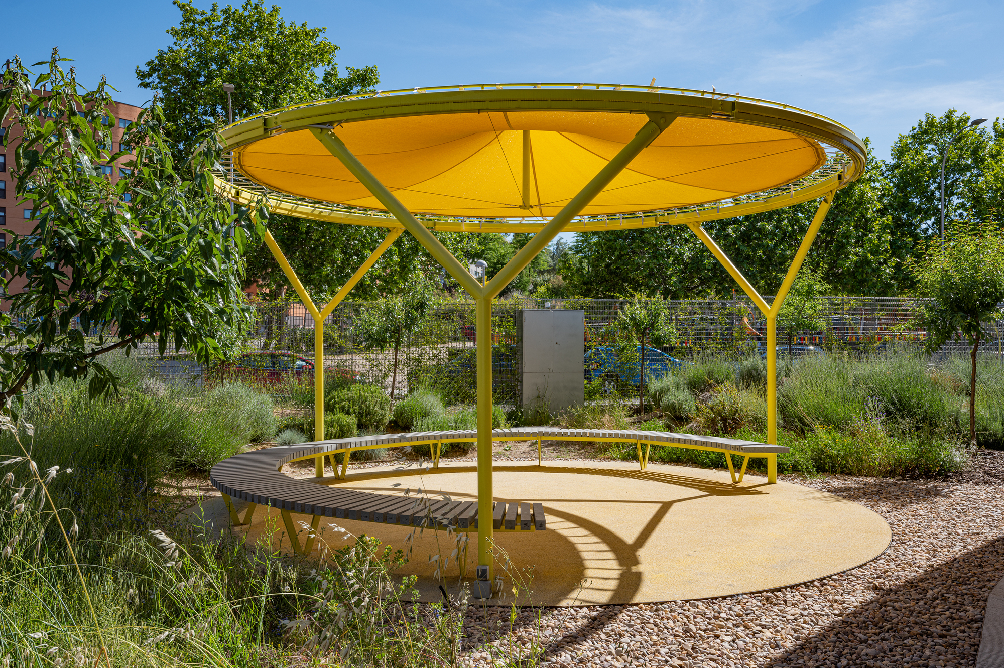 Round yellow fabric canopy on slender steel legs above a curved slatted bench and circular yellow floor, ringed by grasses and gravel