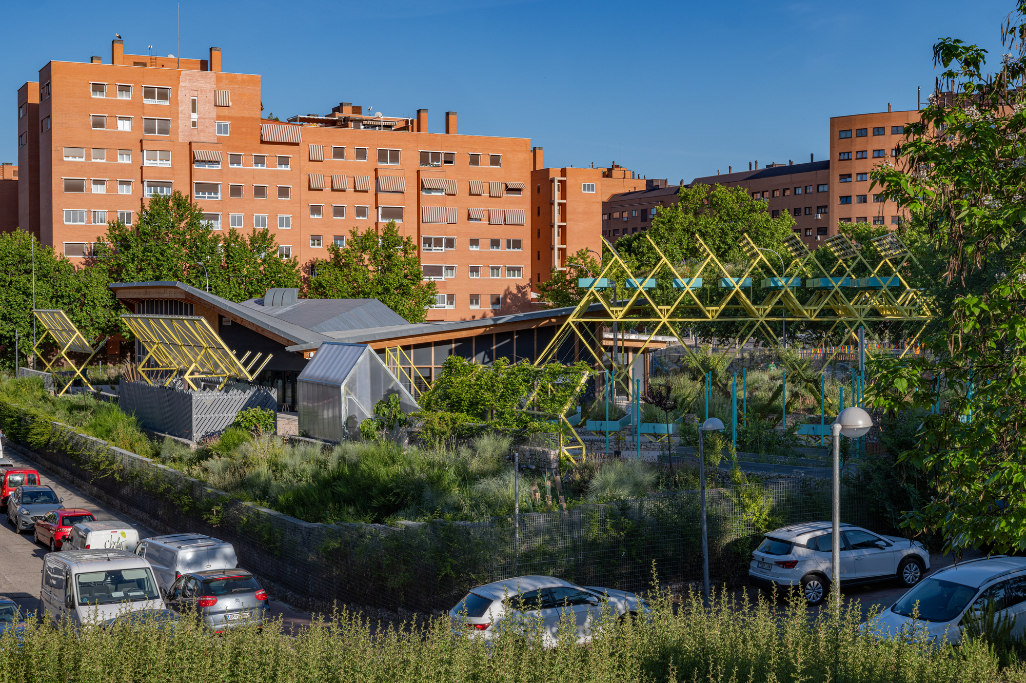 Low timber-roofed pavilion and yellow steel frames behind a planted gabion wall, with brick apartment blocks under a clear blue sky