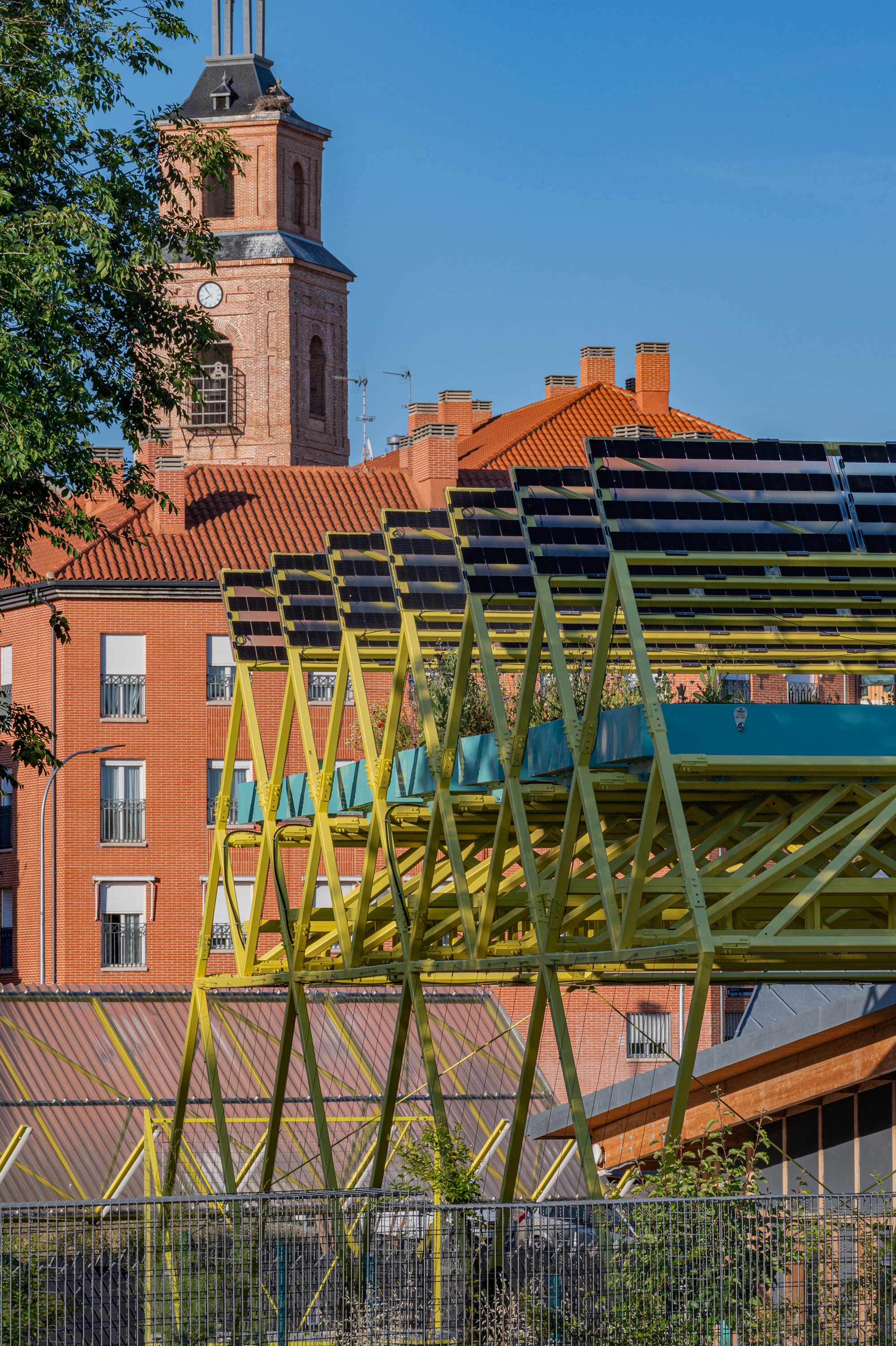 Yellow steel truss carrying solar panels in front of a brick church tower and tiled roofs in late sun