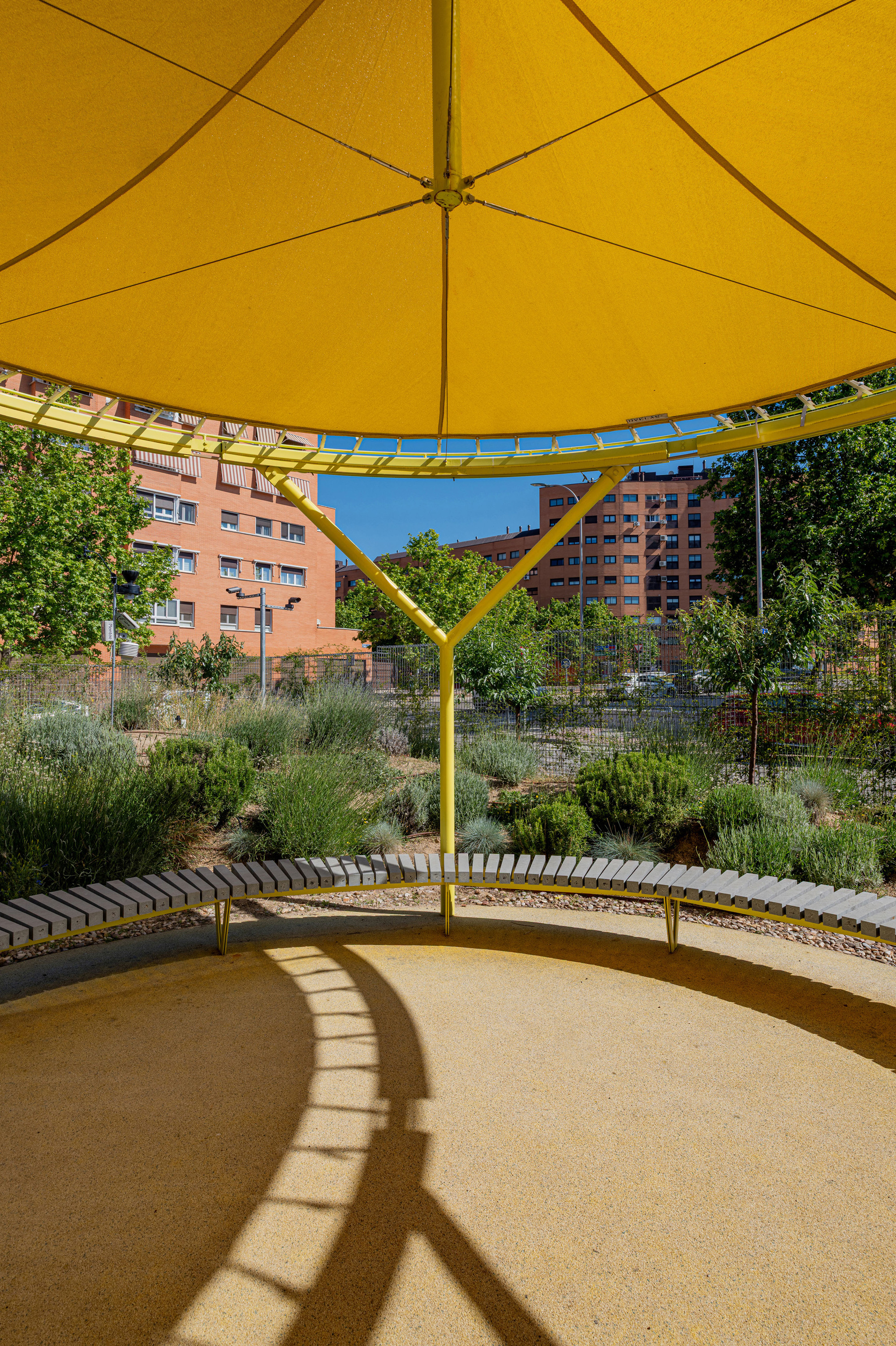 Underside of a yellow fabric canopy over a curved slatted bench and gravel planting, with sunlight casting arched shadows on the ground