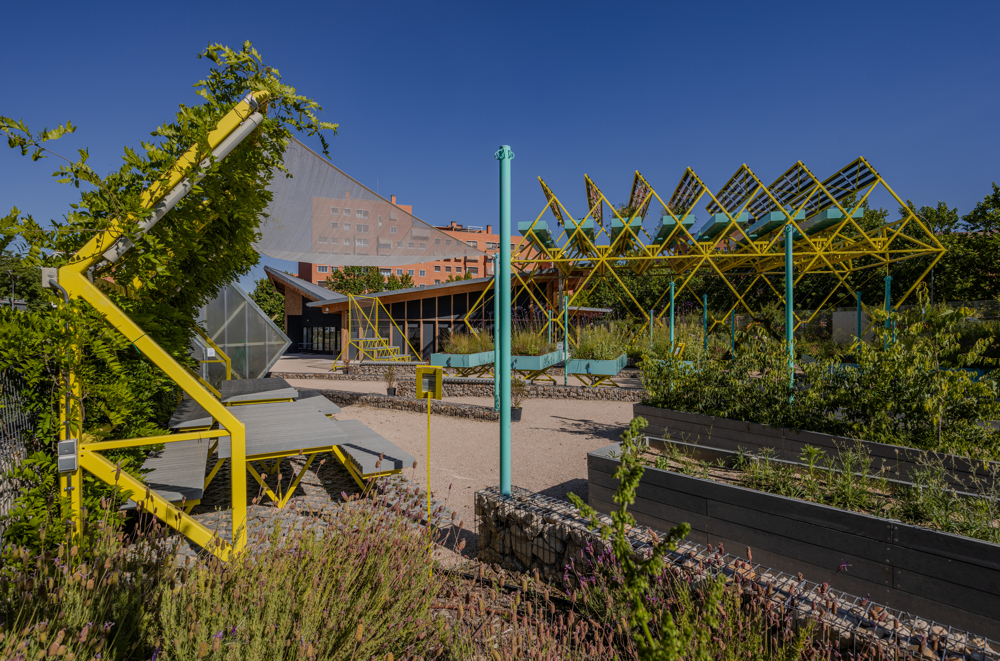 Yellow steel pergola and teal-topped planters across a gravel courtyard, with a vine-covered frame in the foreground against a deep blue sky