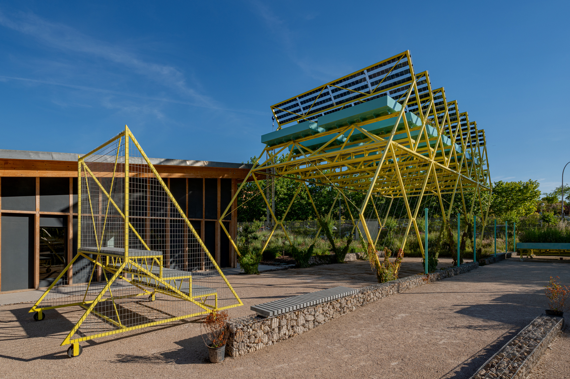 Zigzag yellow steel frame holding solar panels beside a wheeled triangular mesh stair, with a gabion bench in afternoon light