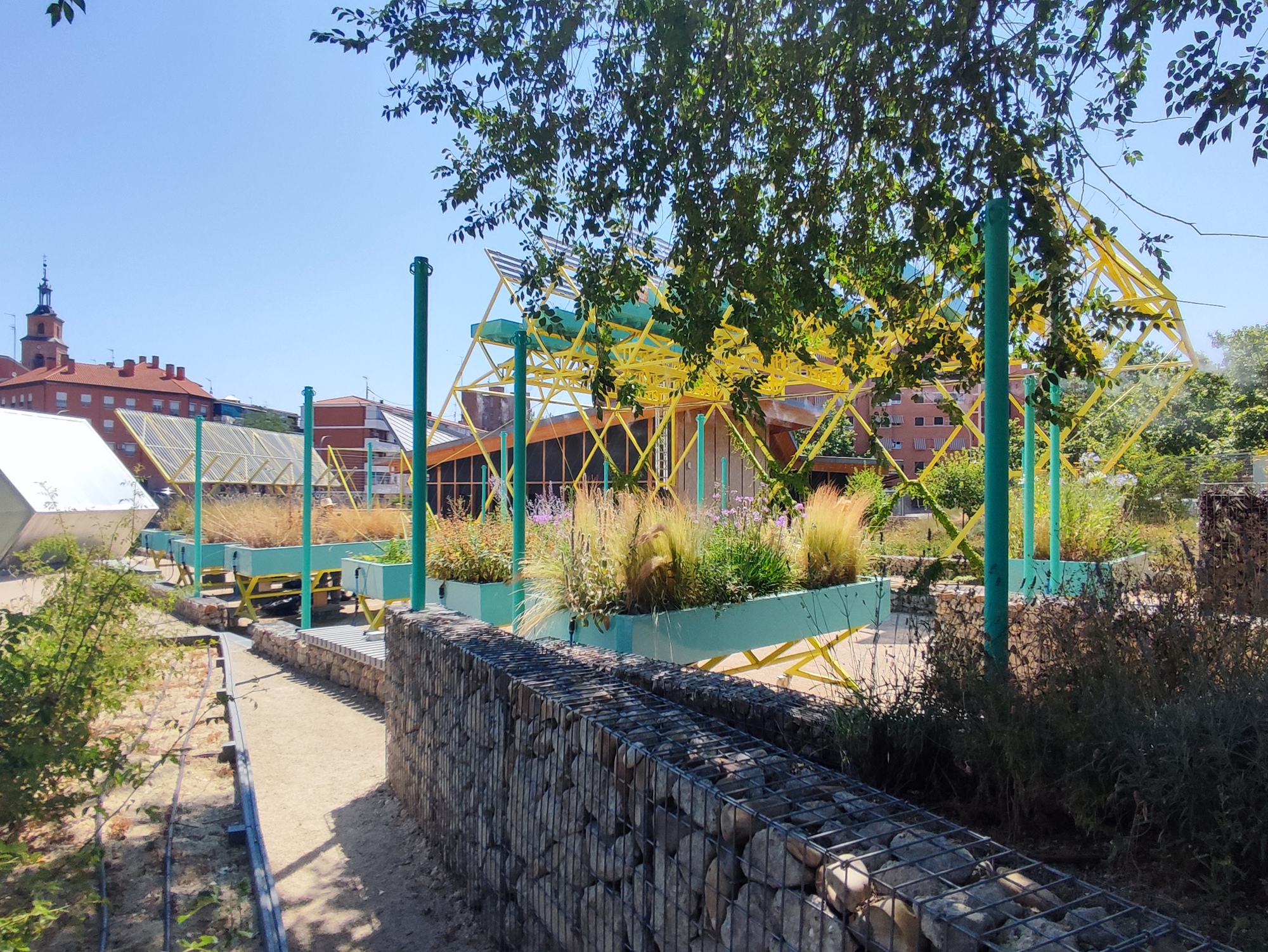 Yellow steel pergola frame beyond teal planter boxes and a gabion stone wall under a clear summer sky