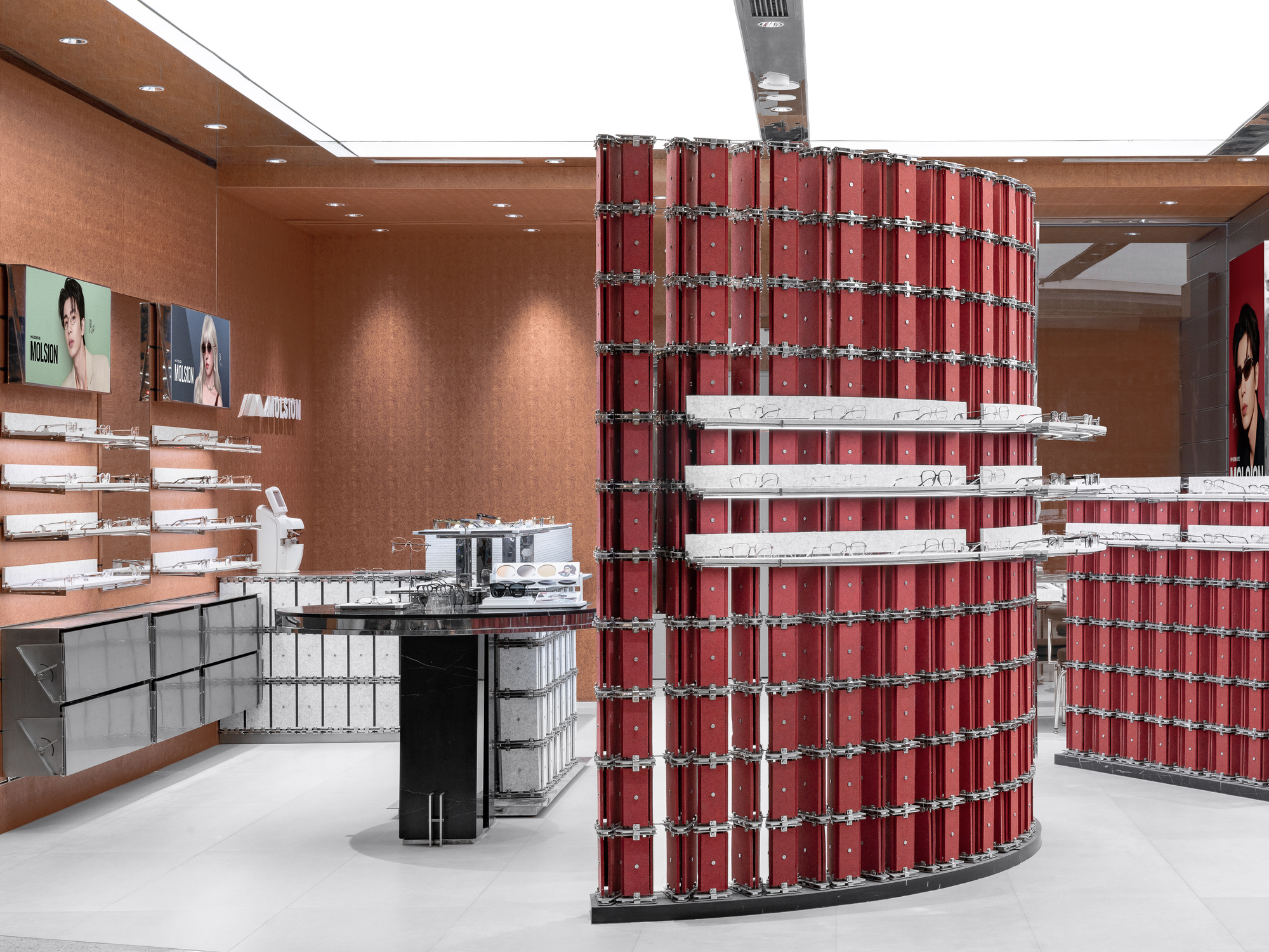 Curved wall of stacked red boxes with chrome clasps and glass shelves holding eyeglasses, set against terracotta-toned walls under a bright white ceiling