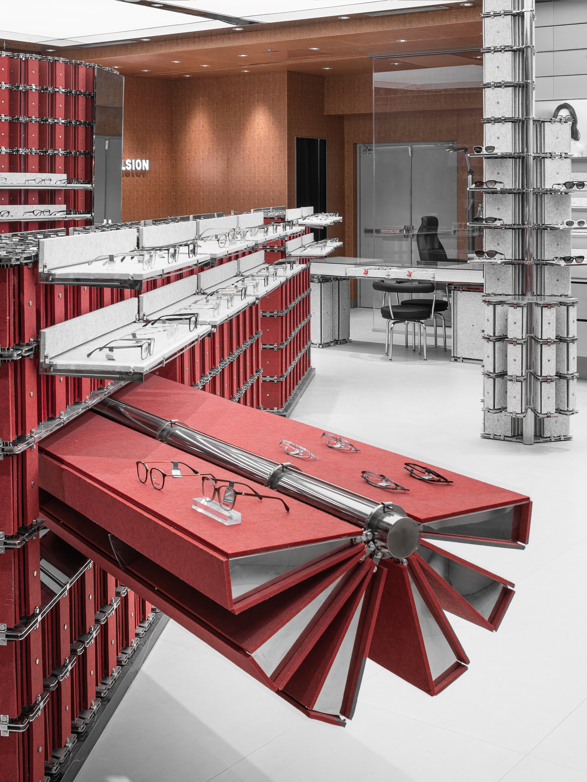 Pleated red display tray cantilevered from a stacked-box wall, holding eyeglasses beside a grey felt-clad column with chrome fittings