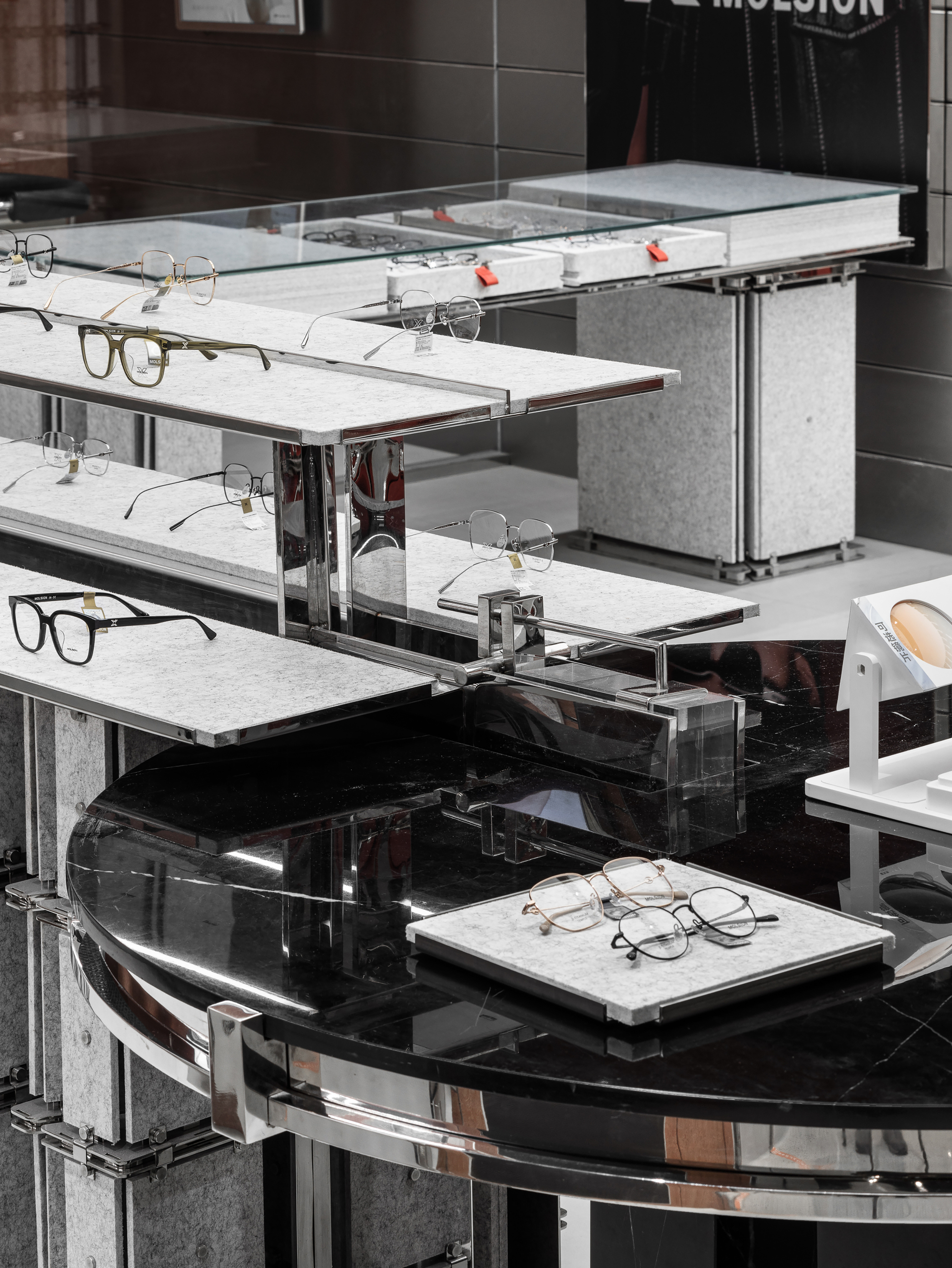 Close view of felt-lined shelves, chrome brackets and a black marble round table displaying eyeglass frames, with a glass-topped case behind