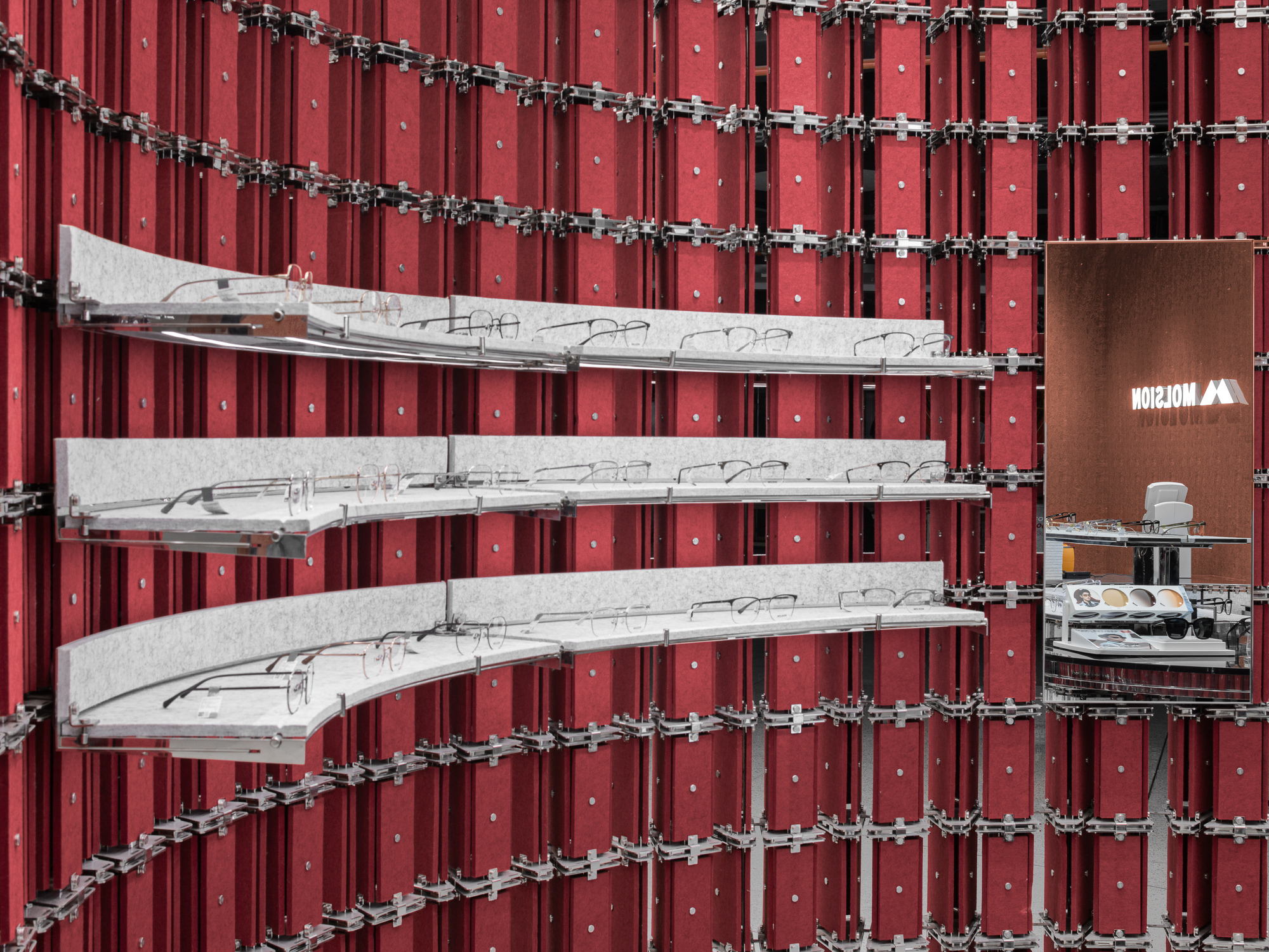 Curved shelves of eyeglasses projecting from a wall of red felt panels joined by steel clamps, with a mirror reflecting the store