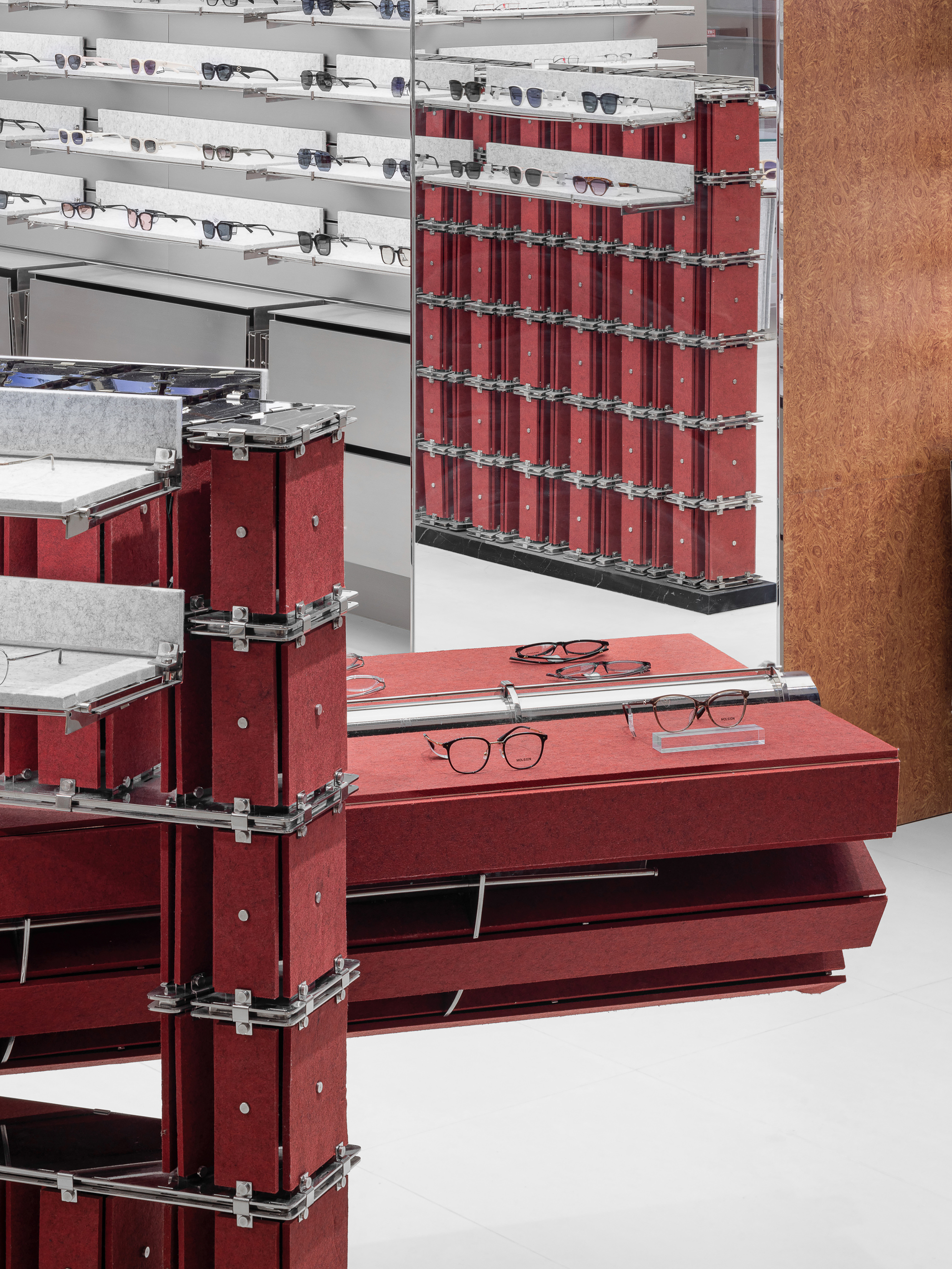 Close-up of red felt display blocks bolted with steel brackets, glasses resting on a cantilevered slab beside a mirror