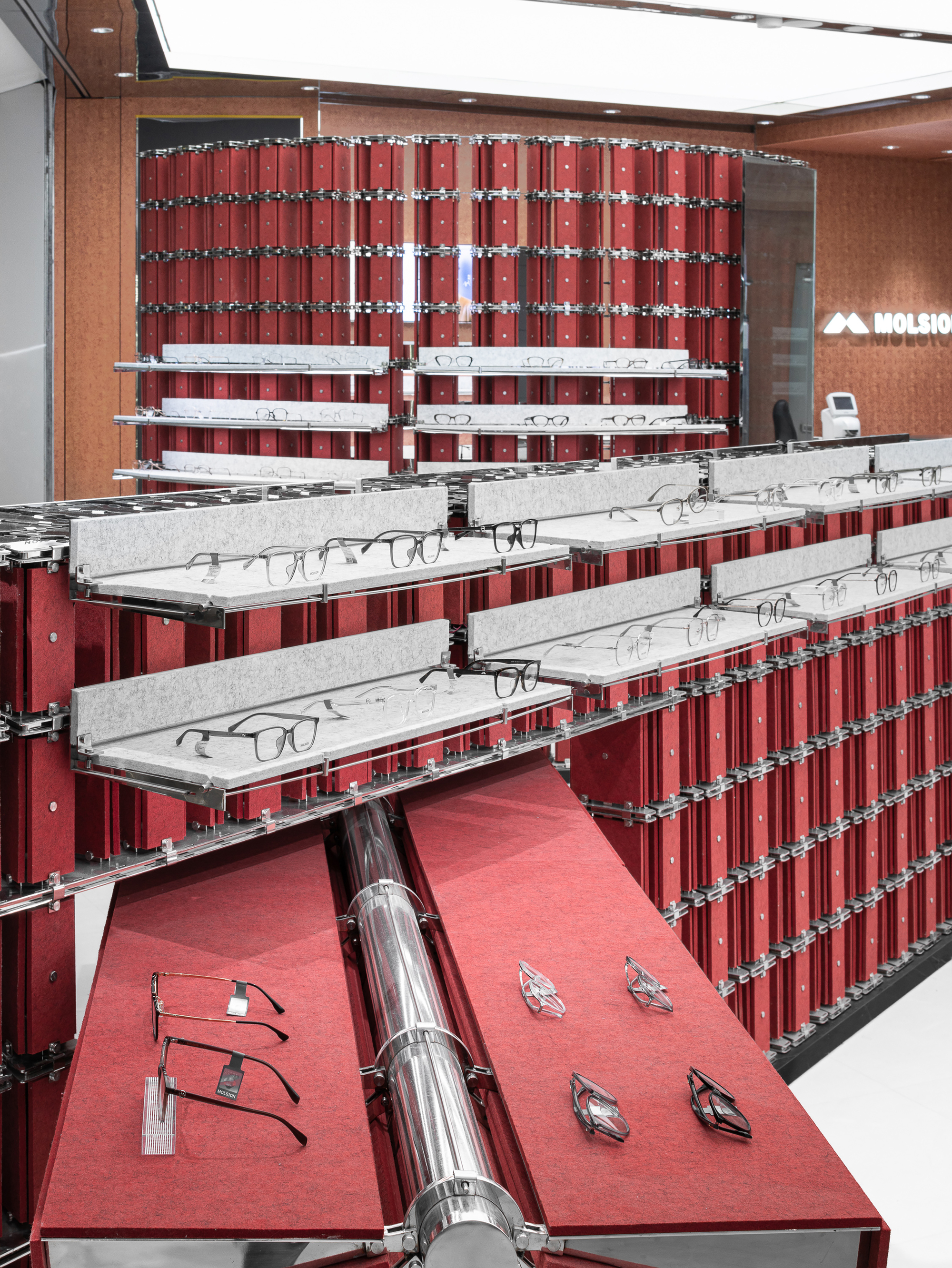 Tiered glasses shelves on a red felt wall, with a polished steel cylinder crossing red display tops in the foreground