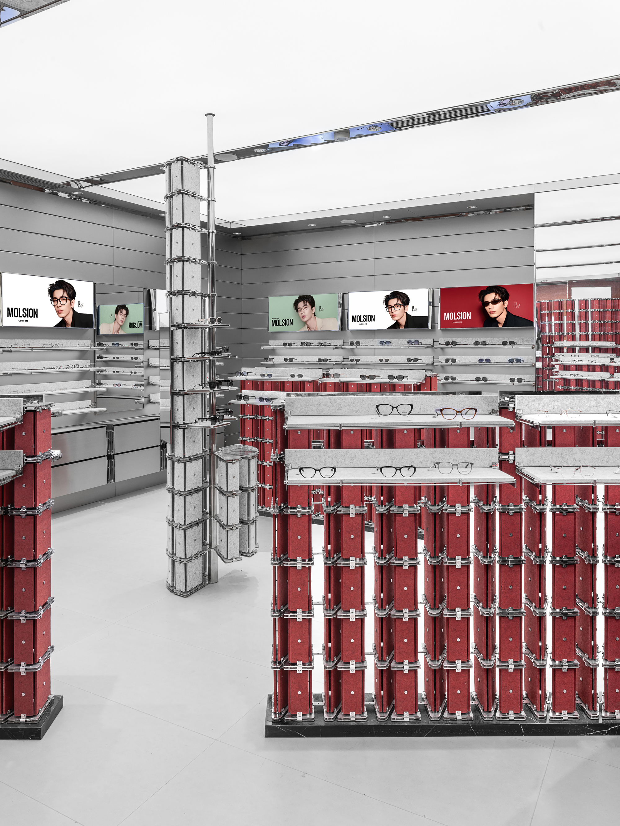 Rows of red felt-panel display stands holding eyeglasses, with a slim grey column and portrait light boxes on the back wall