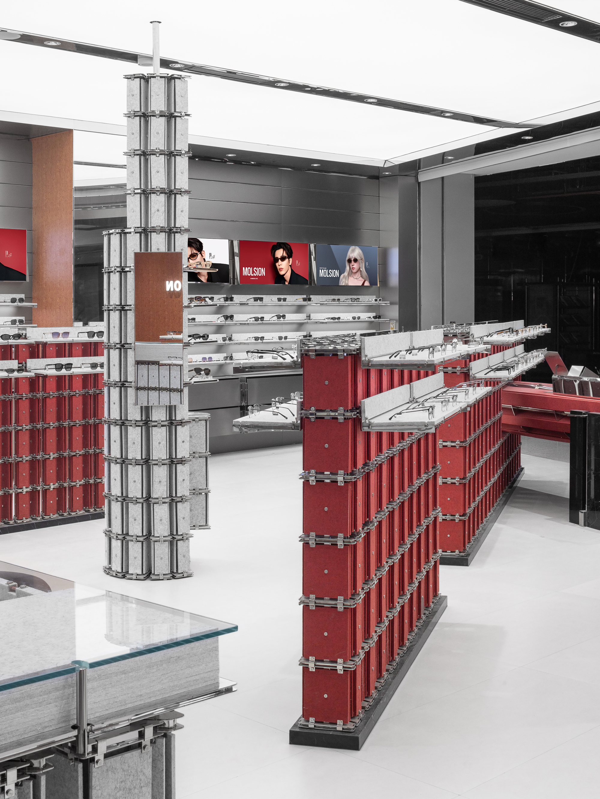 Red display wall with angled steel shelves beside a grey panel column and a glass-topped table in the foreground