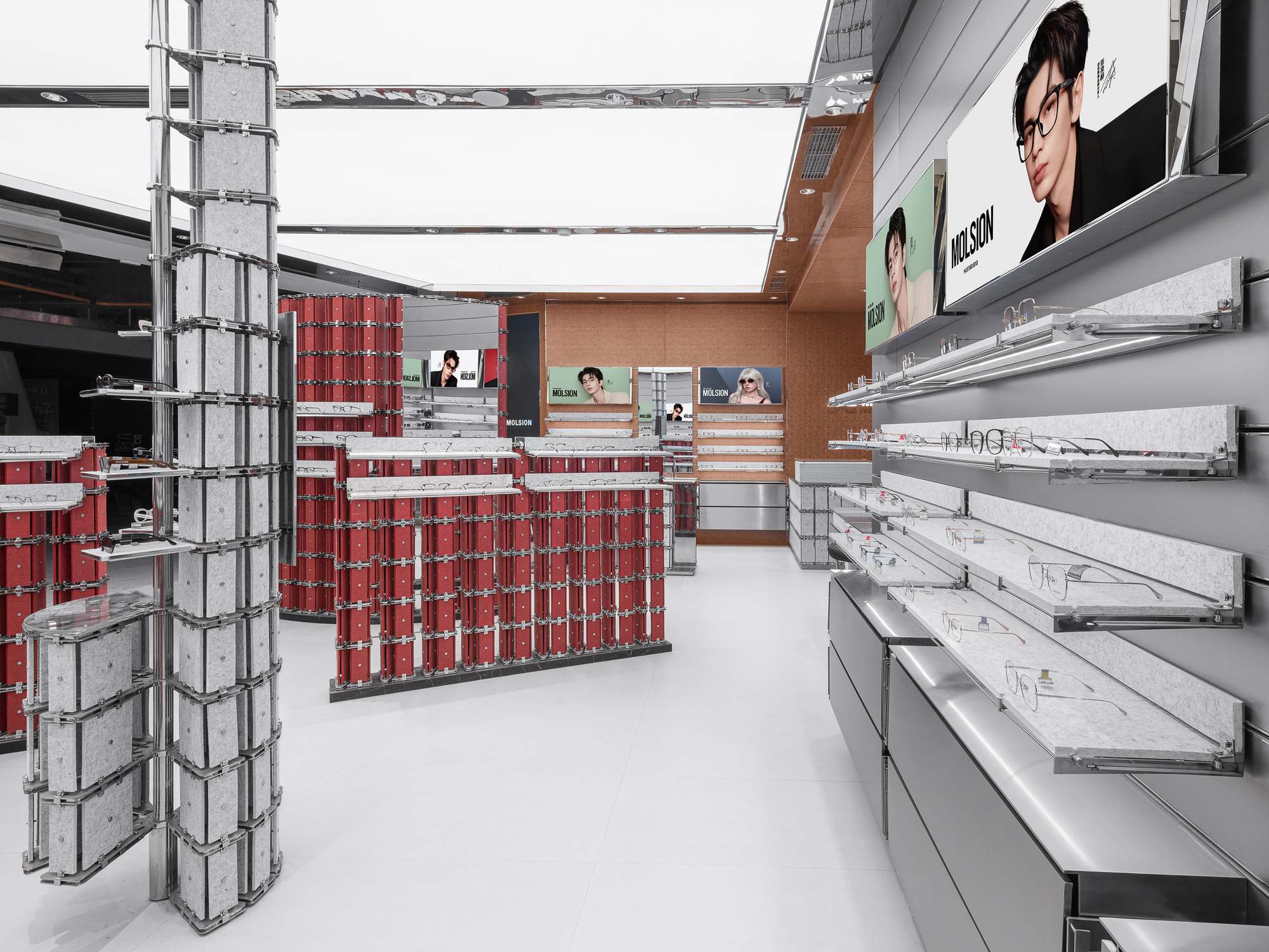 Aisle lined with steel wall shelves of eyeglasses, leading toward red display stands and a warm timber-toned back wall