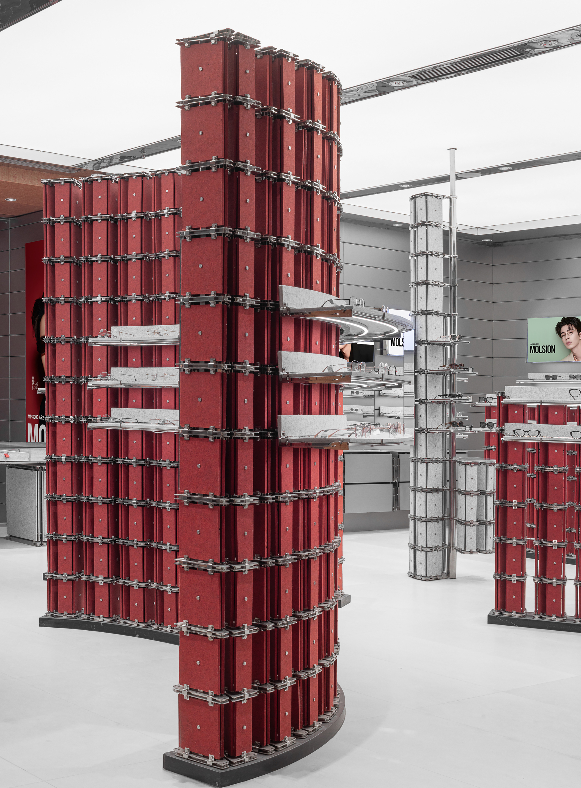 Curved red display wall of clamped felt panels with cantilevered shelves, set against a grey column and a luminous ceiling