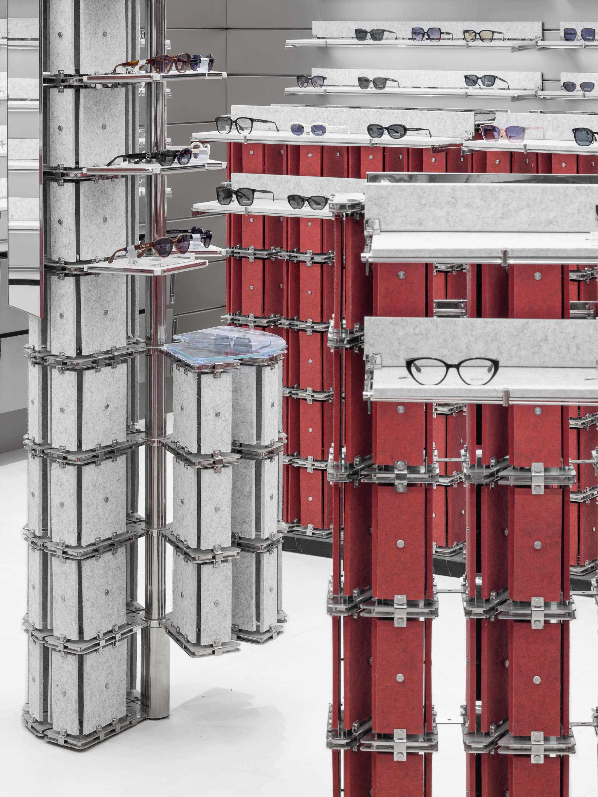 Stacked red and grey felt panel columns clamped with steel brackets, holding sunglasses on glass-topped shelves above a white floor