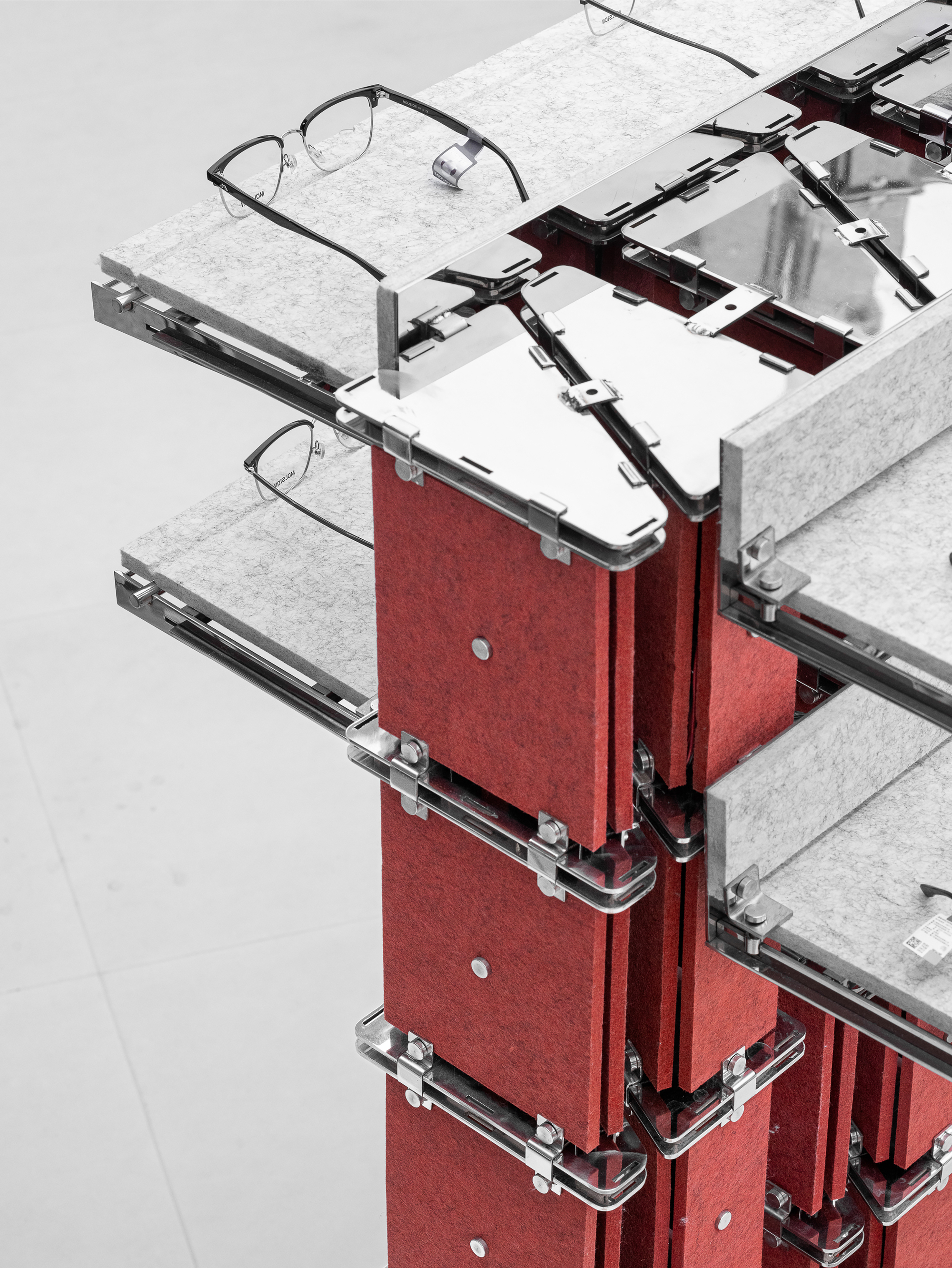 Corner detail of a red felt column with clear acrylic clamps and mirrored steel trays holding eyeglasses on a pale floor