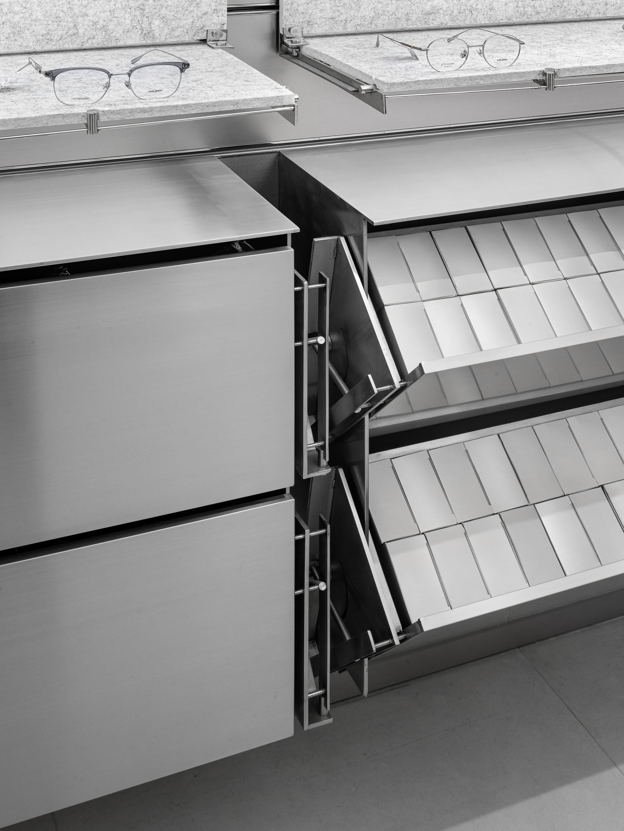 Brushed steel drawers with one pulled open to show tilted white trays, with eyeglasses on felt shelves above