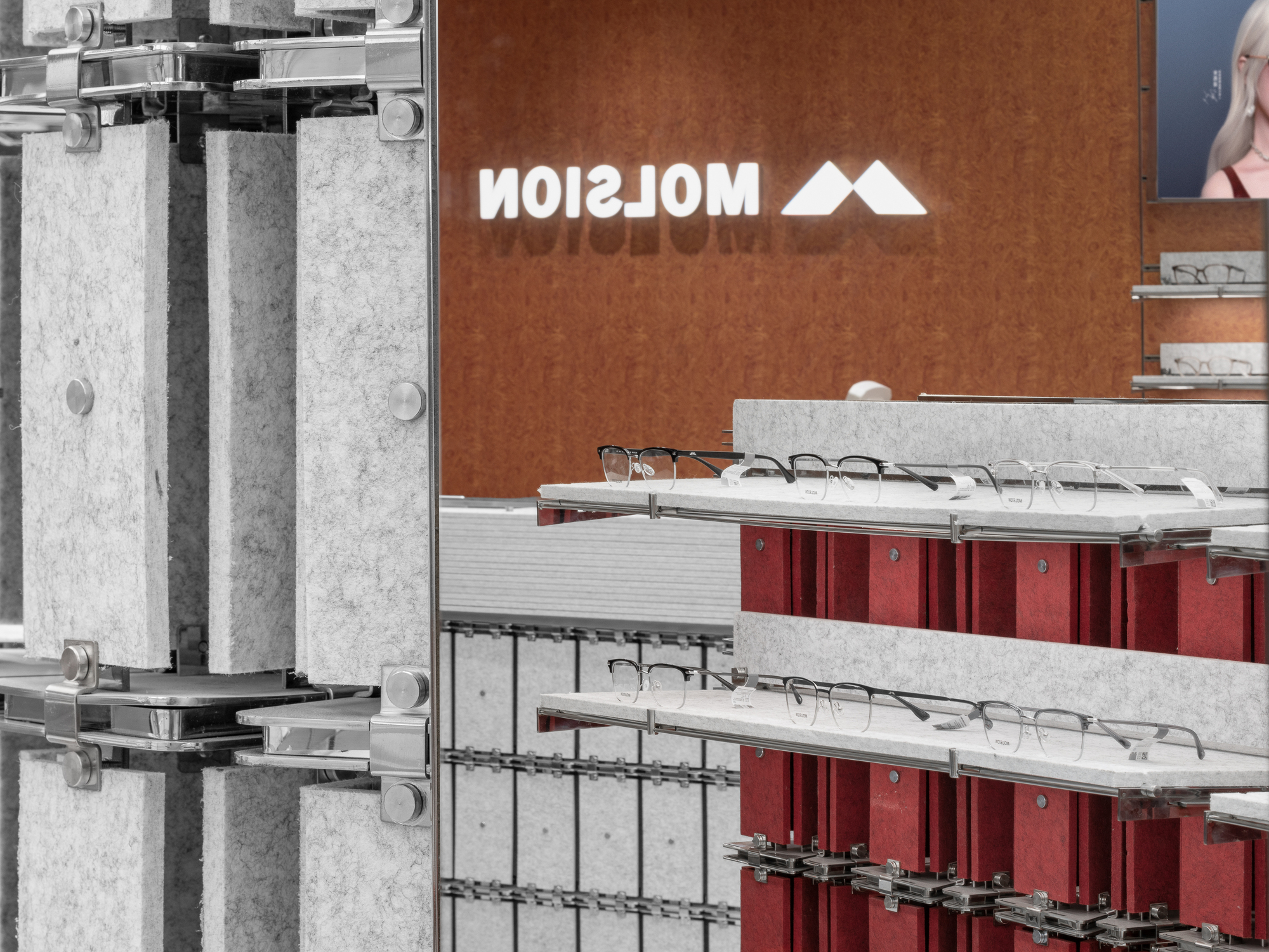 Glowing mirrored logo on a burl wood wall, seen past grey felt columns and red-backed shelves lined with eyeglasses
