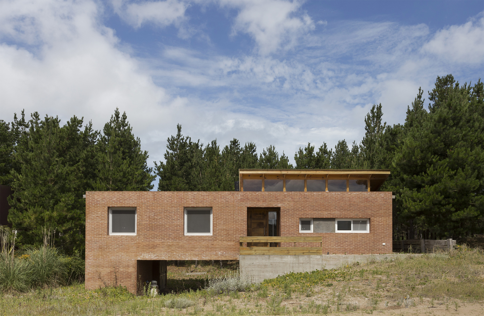 Red brick house elevated on a concrete base with a clerestory roof, set against pine trees under a partly cloudy sky