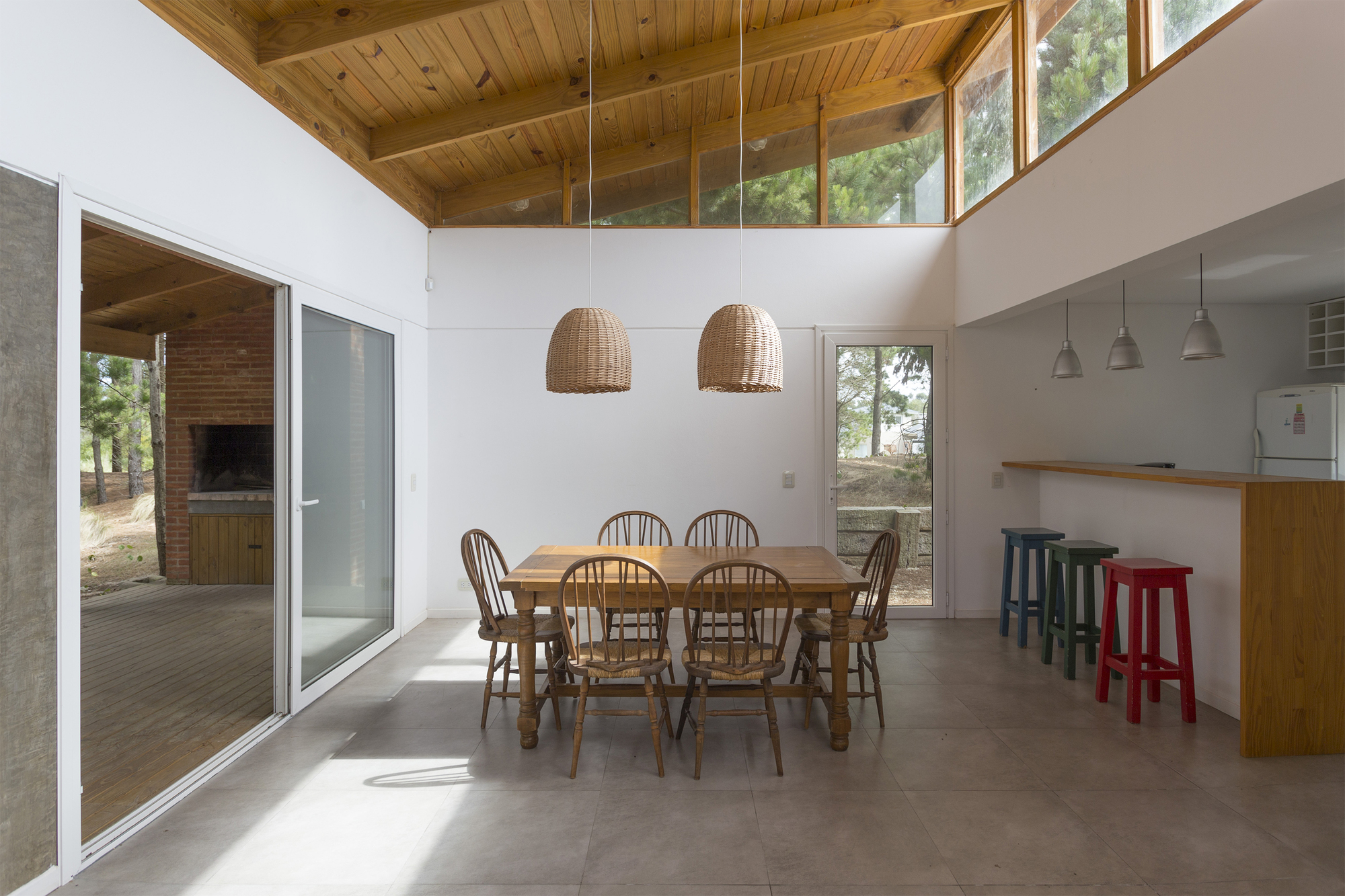 Dining area with wooden table, spindle chairs and wicker pendants beneath a pitched timber ceiling, sunlight falling across the tile floor