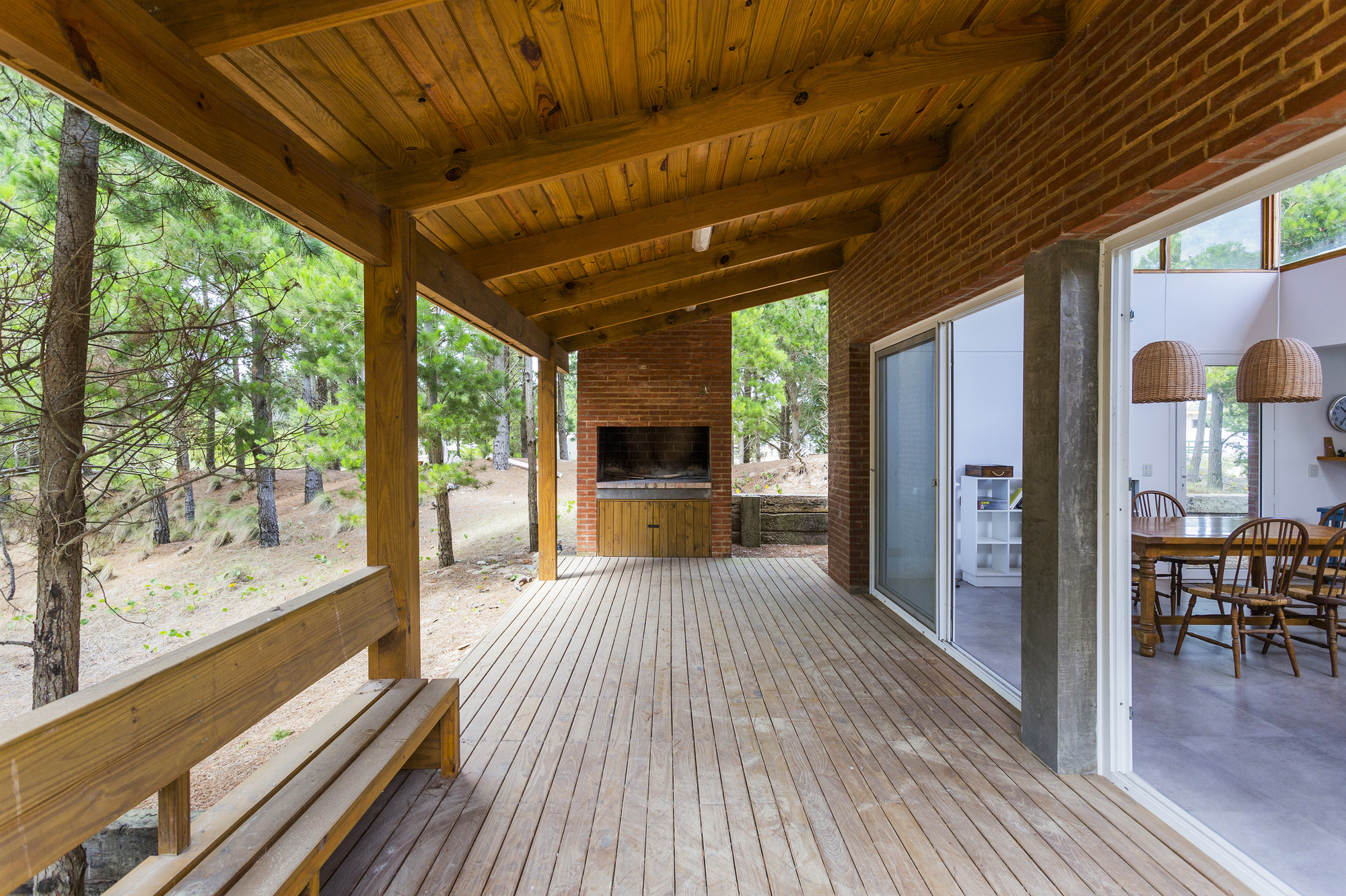 Covered timber deck with built-in bench and brick grill, framed by pine trunks and open to the dining room