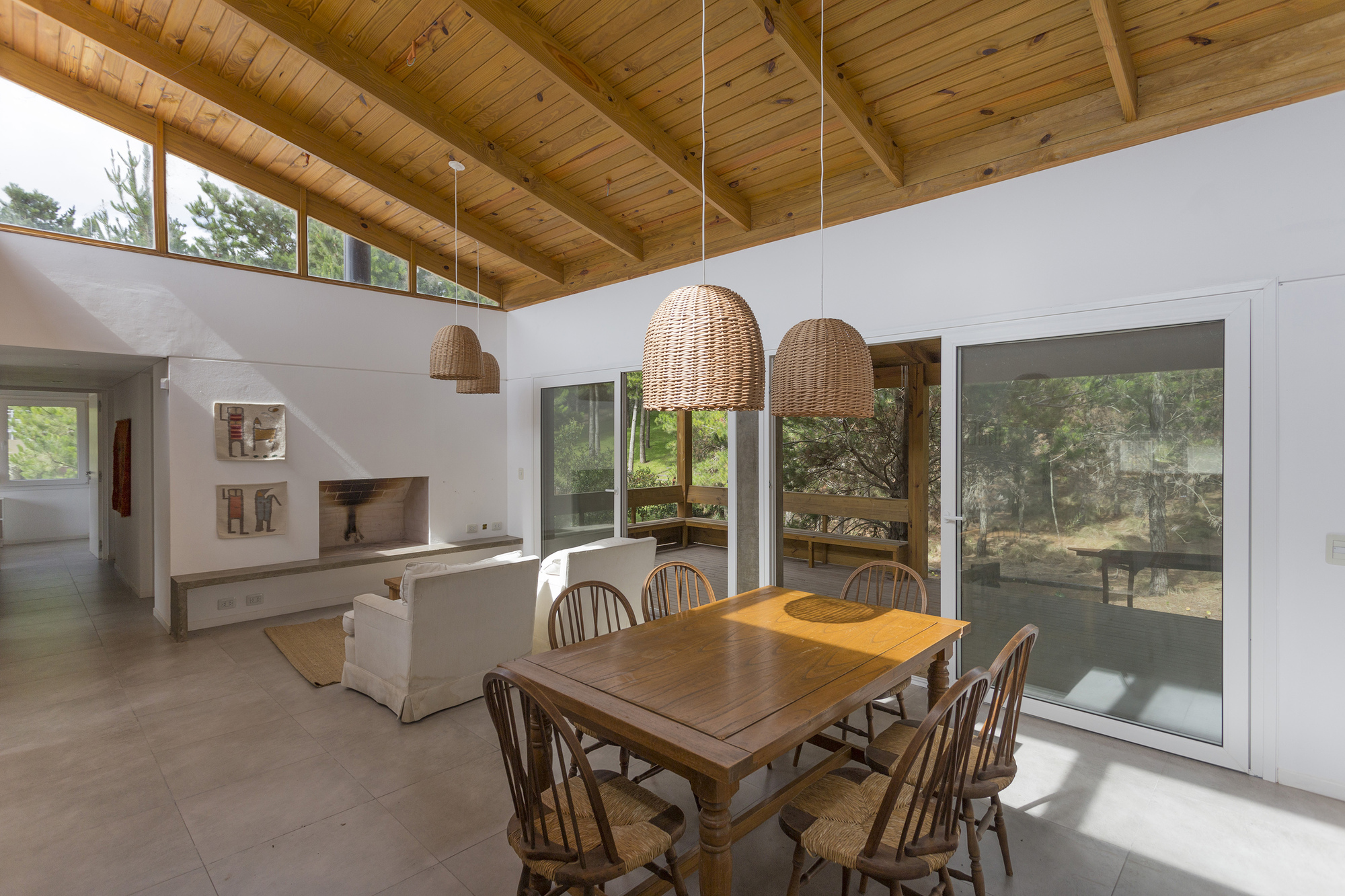 Living and dining space with brick-lined fireplace, white armchairs and wicker pendants, daylight entering through clerestory windows and sliding glass doors