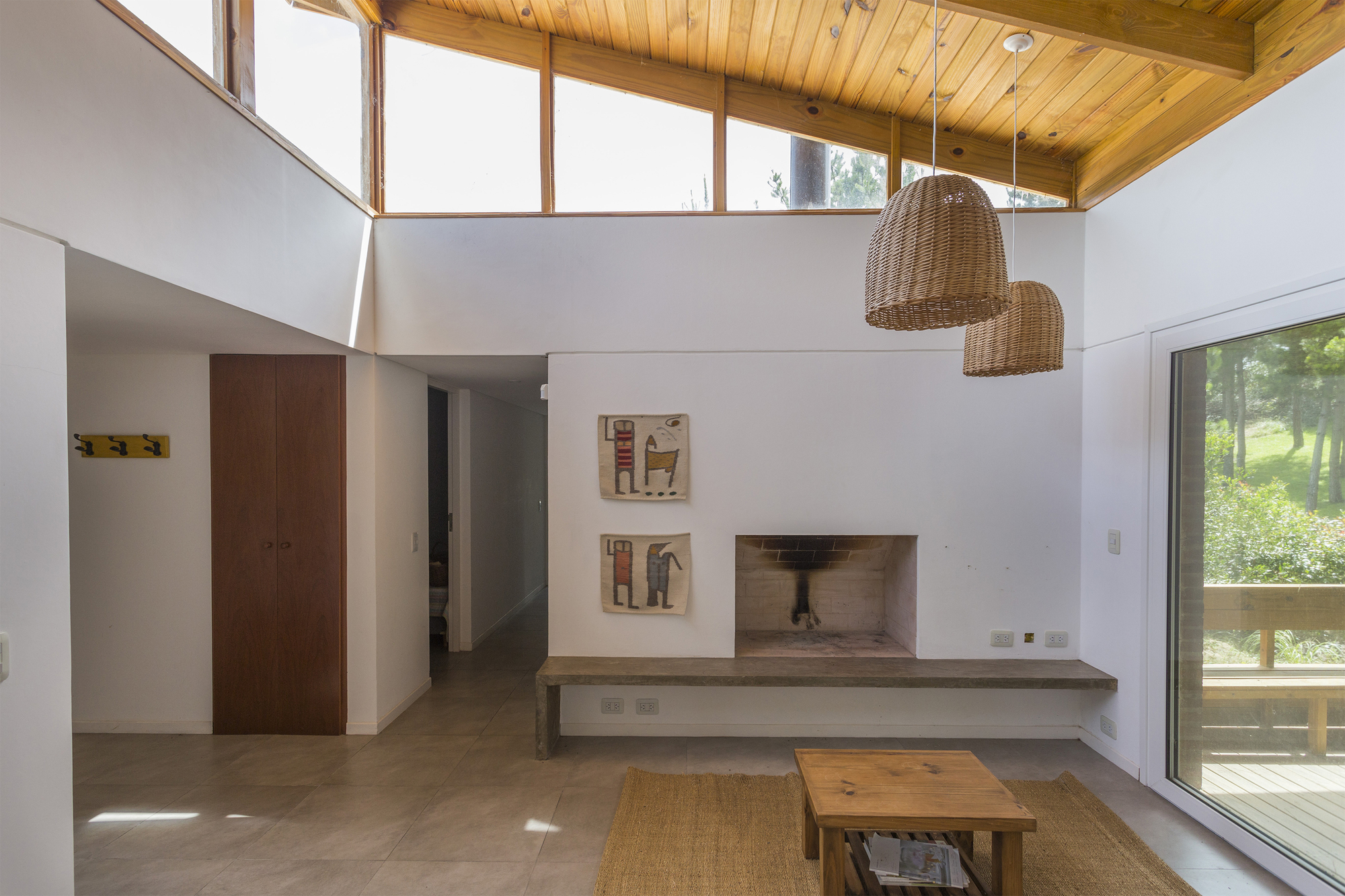 Living room with a recessed brick fireplace, concrete bench and wicker pendants beneath a timber clerestory ceiling, with a deck visible through the sliding gla