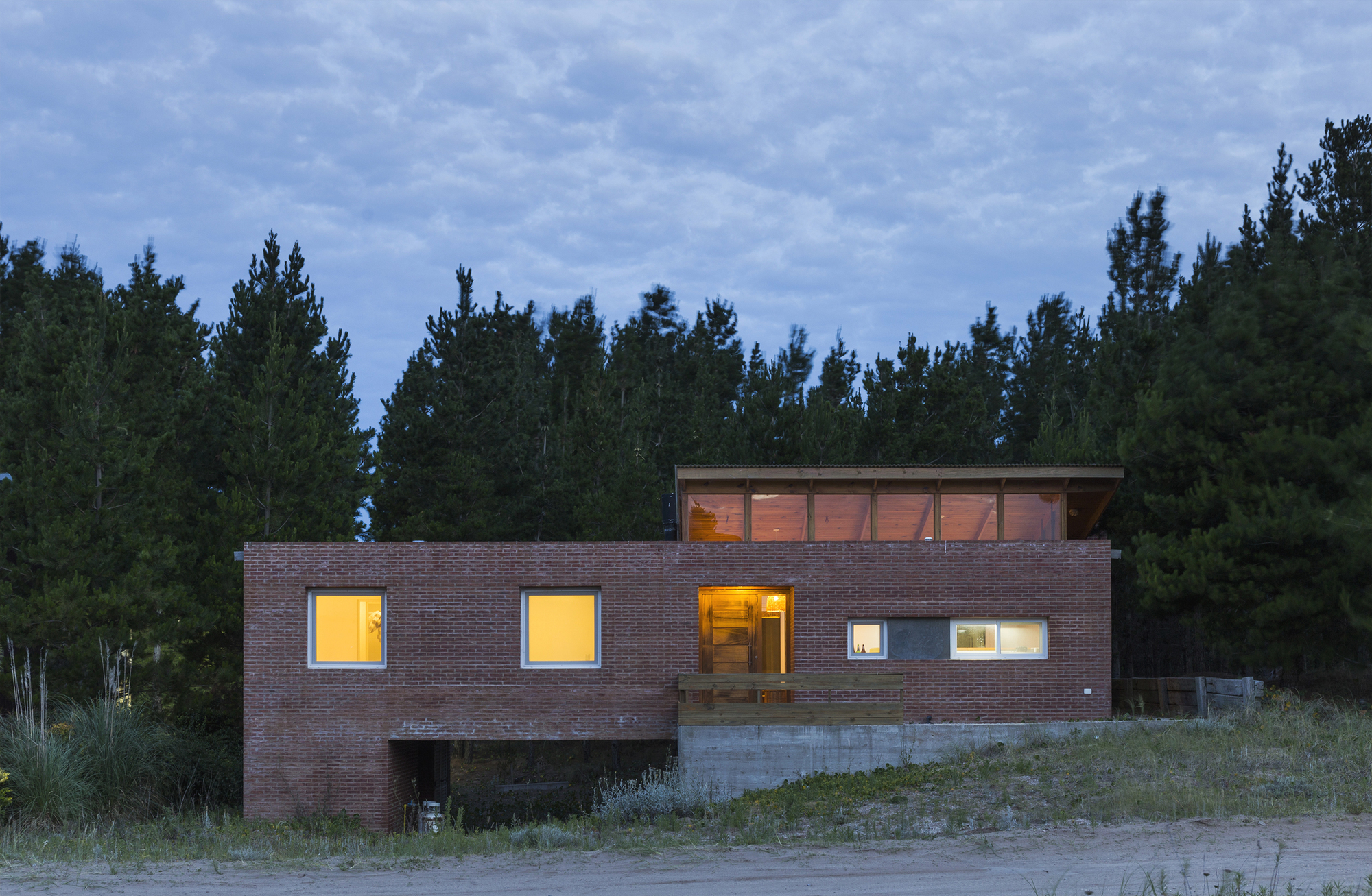 Brick house with lit windows and a glazed timber clerestory, set against a dark pine stand at dusk