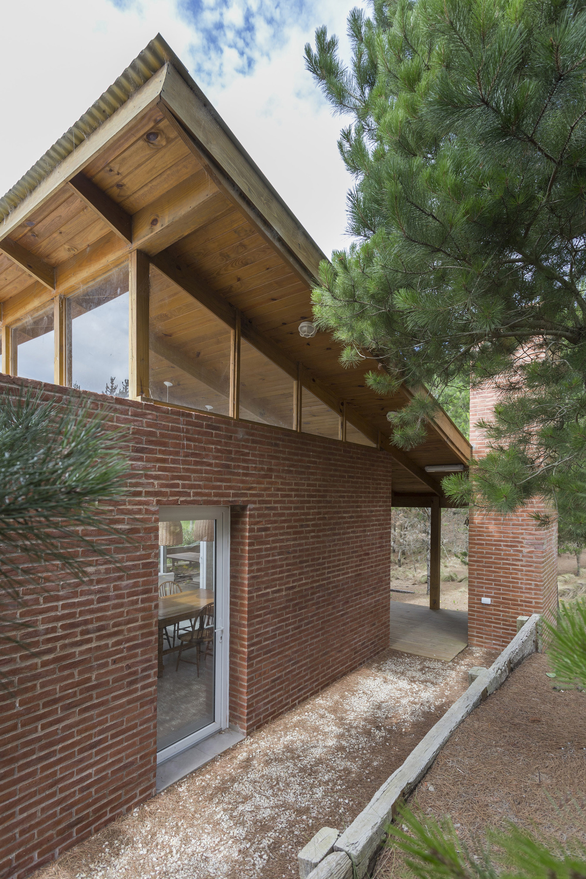 Side wall of red brick with a glazed door beneath a sloped timber roof, framed by pine branches