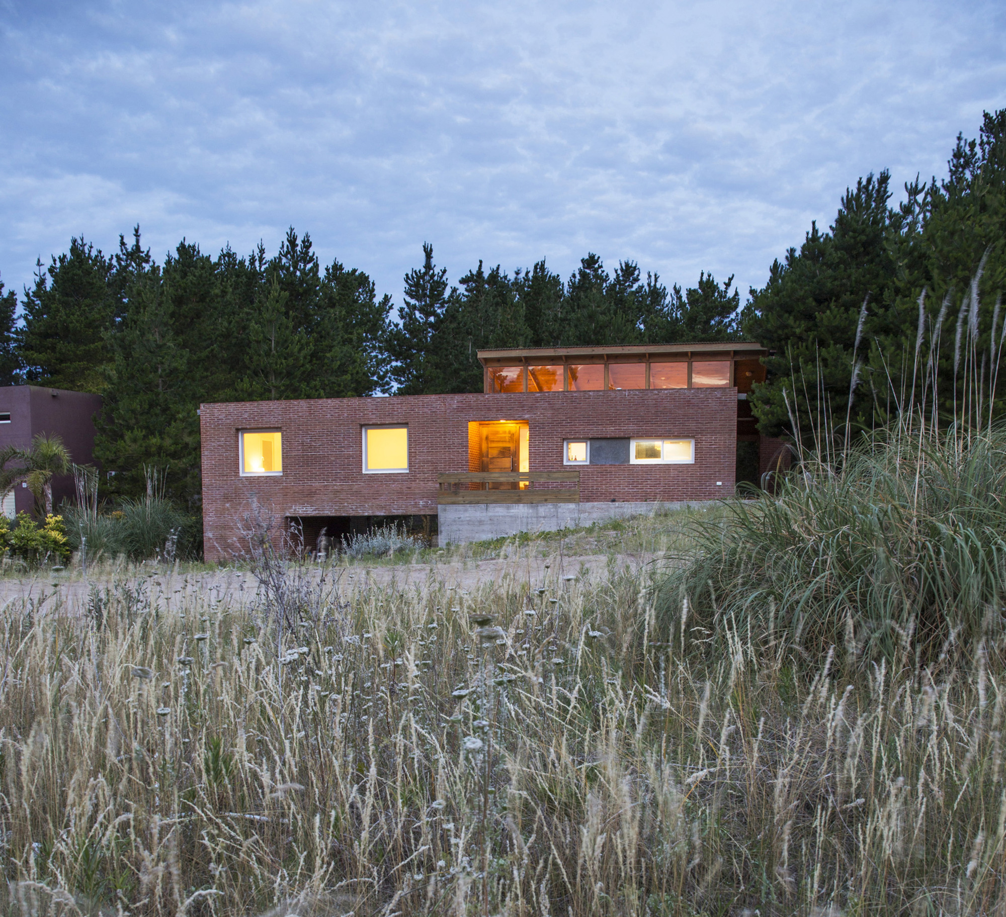 Lit brick house with a wooden entry deck and clerestory, viewed through dune grasses under a cloudy dusk sky
