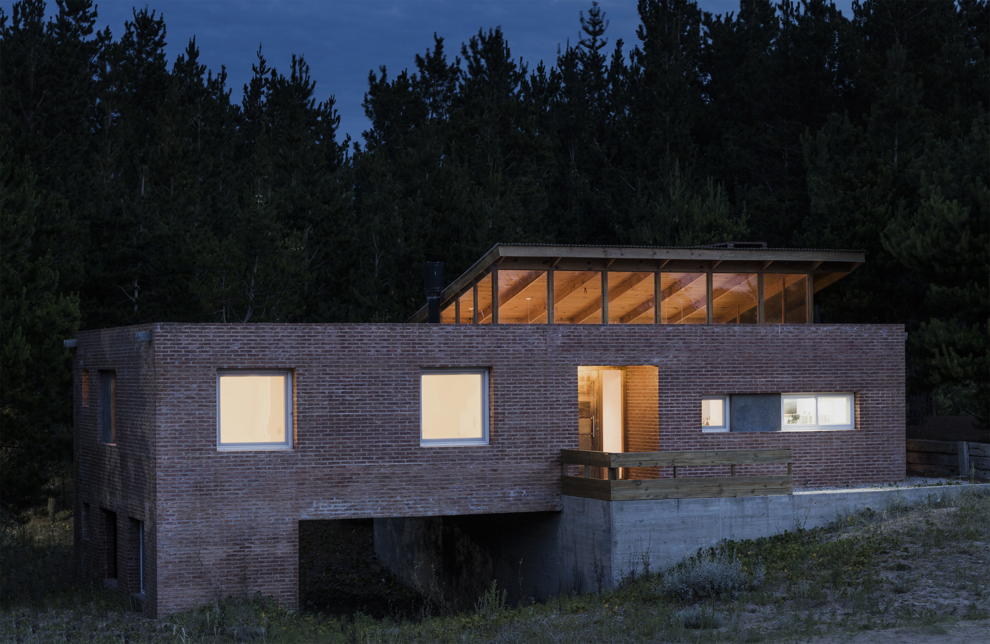 Brick volume with glowing windows and a timber-framed clerestory under a cantilevered roof, against dark pines at night