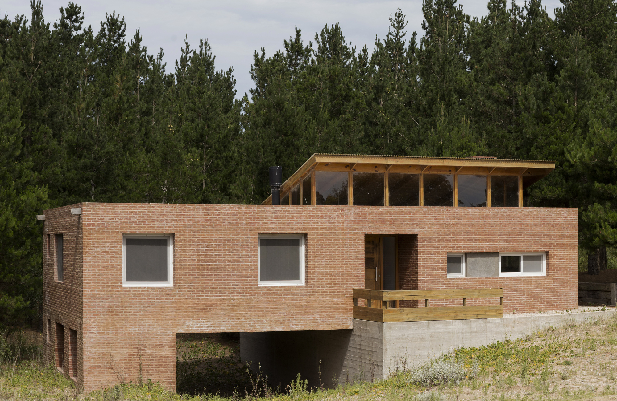 Brick volume with a clerestory of timber-framed glazing under a tilted roof, raised on a concrete base against dense pines