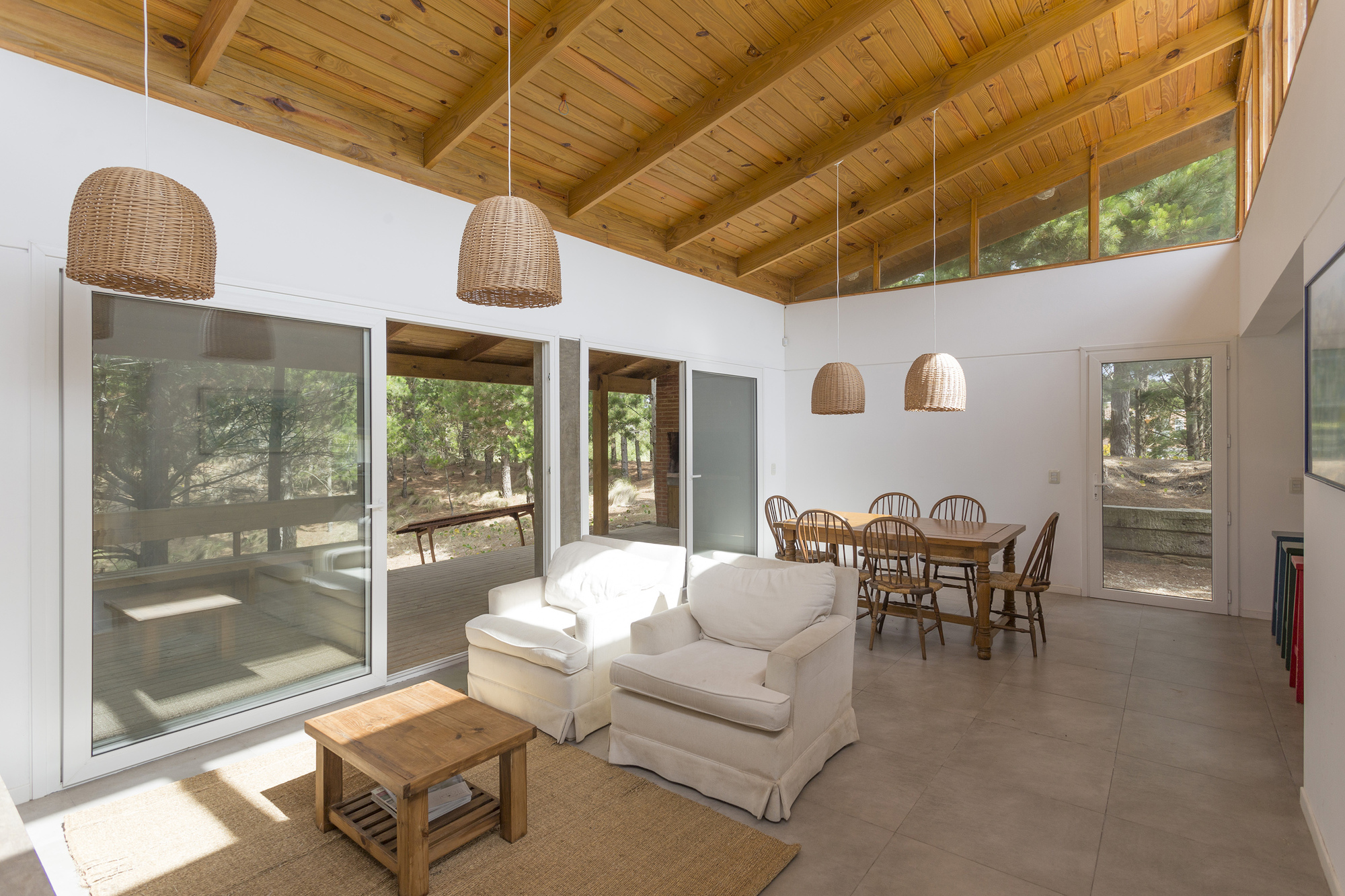 Living and dining room with exposed timber rafters, wicker pendant lamps and sliding glass doors opening to the pine woods