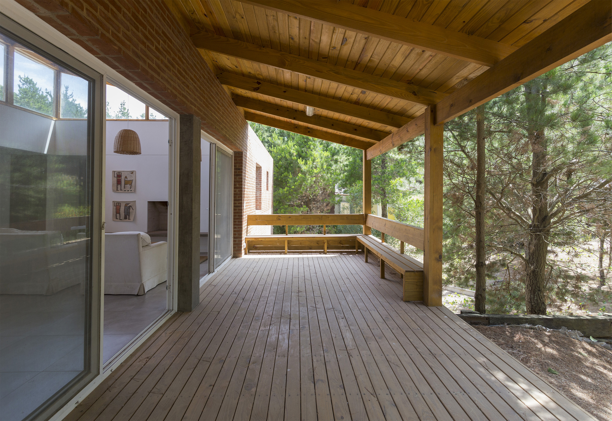 Covered timber deck with built-in bench, heavy beams and plank ceiling, flanked by sliding glass doors and pine trunks