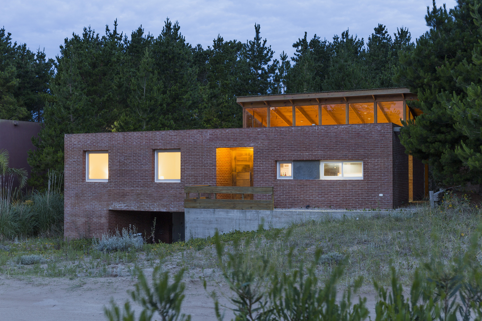Brick facade at dusk with lit windows and a glowing clerestory under the roof, surrounded by pines and dune grass