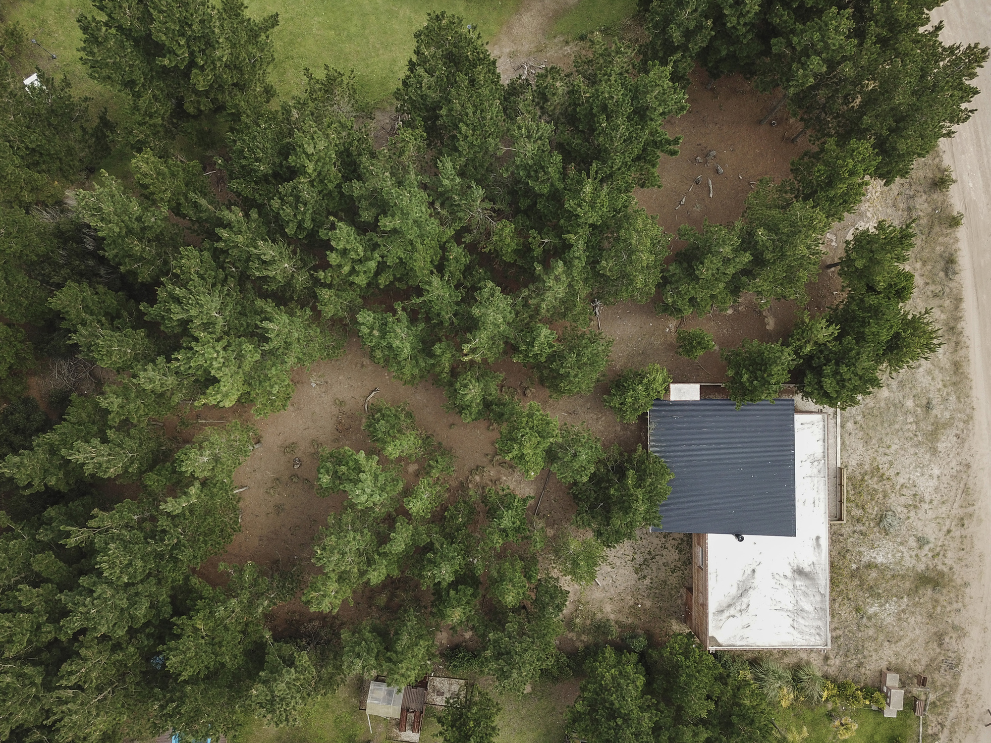 Top-down drone view of the house with dark and white roof planes among pine trees beside a sandy road