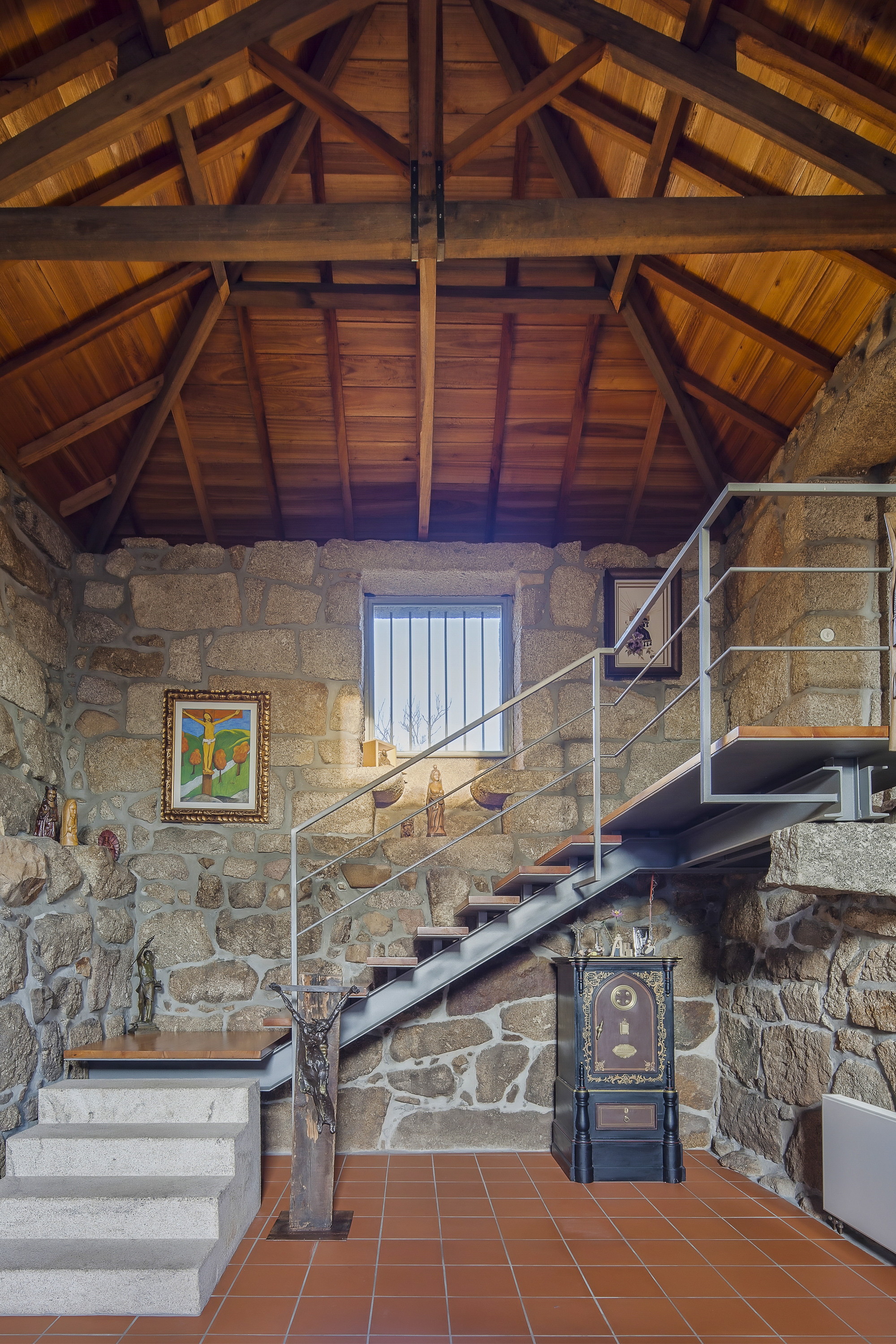 Interior stone wall with a steel stair, timber treads and exposed roof trusses above a terracotta tile floor
