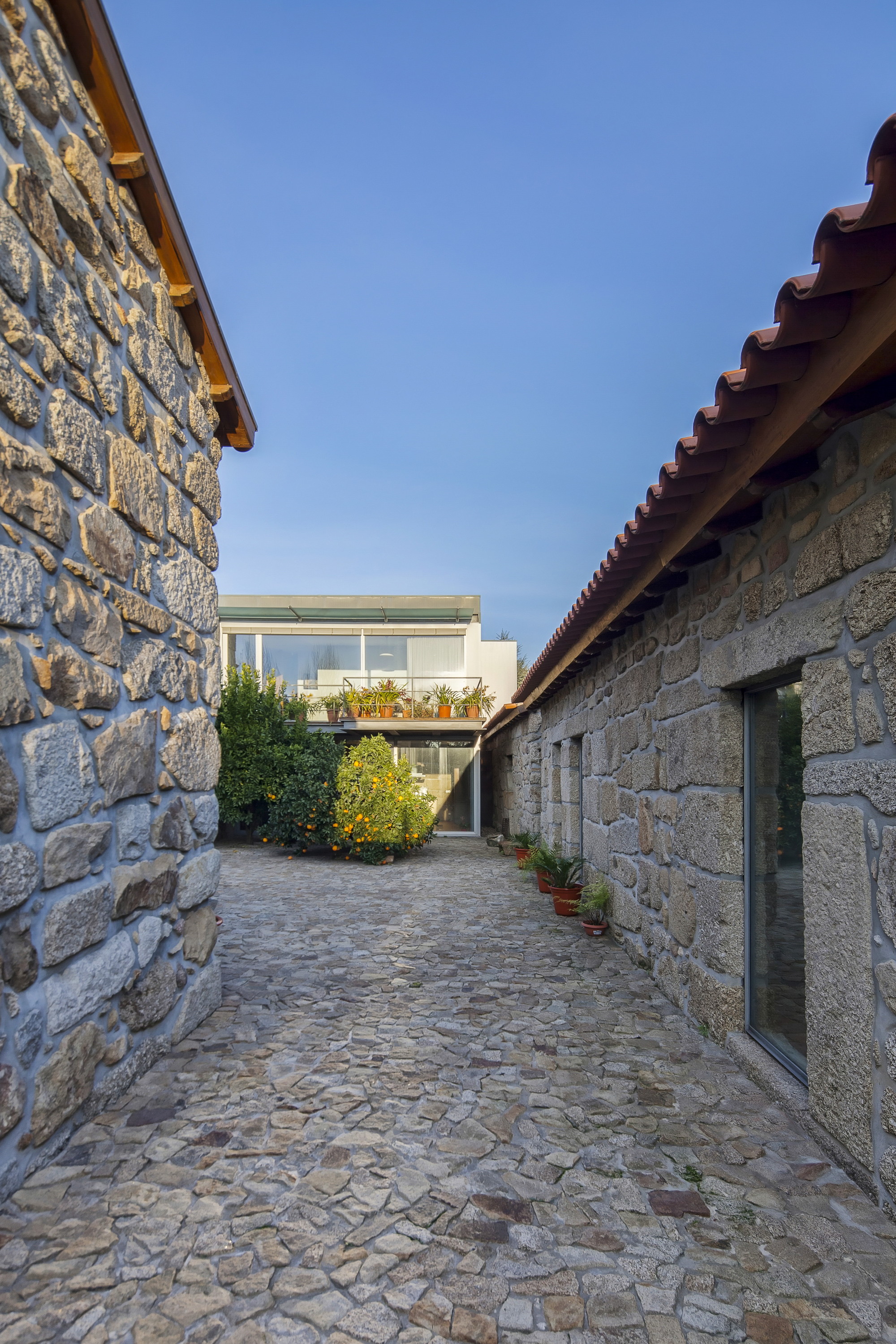 Narrow granite lane between two stone walls, with an orange tree and a glazed upper volume at the far end