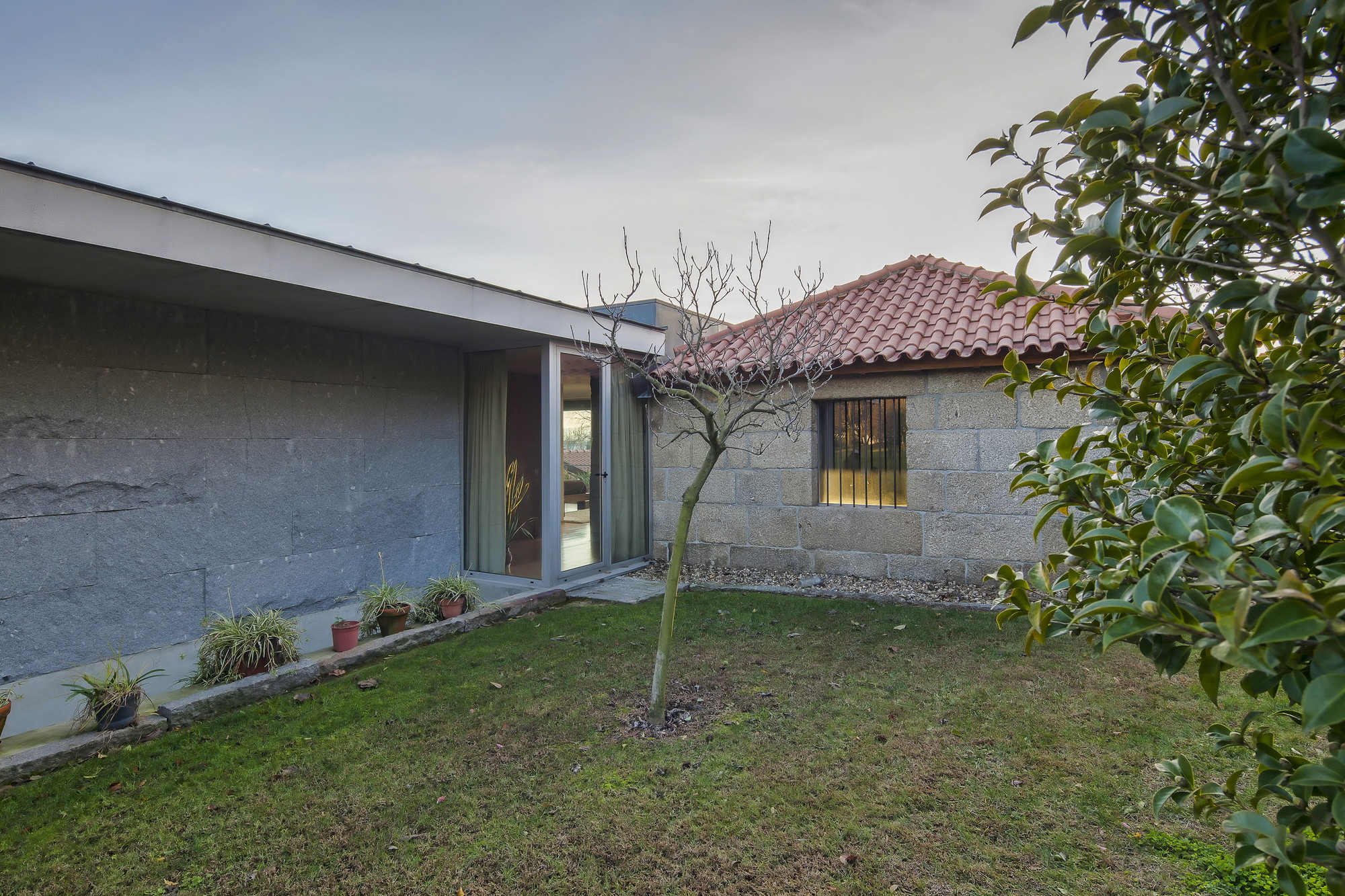 Garden corner where a concrete-edged glass wing meets a granite block wall and tiled roof, with a bare sapling on the lawn