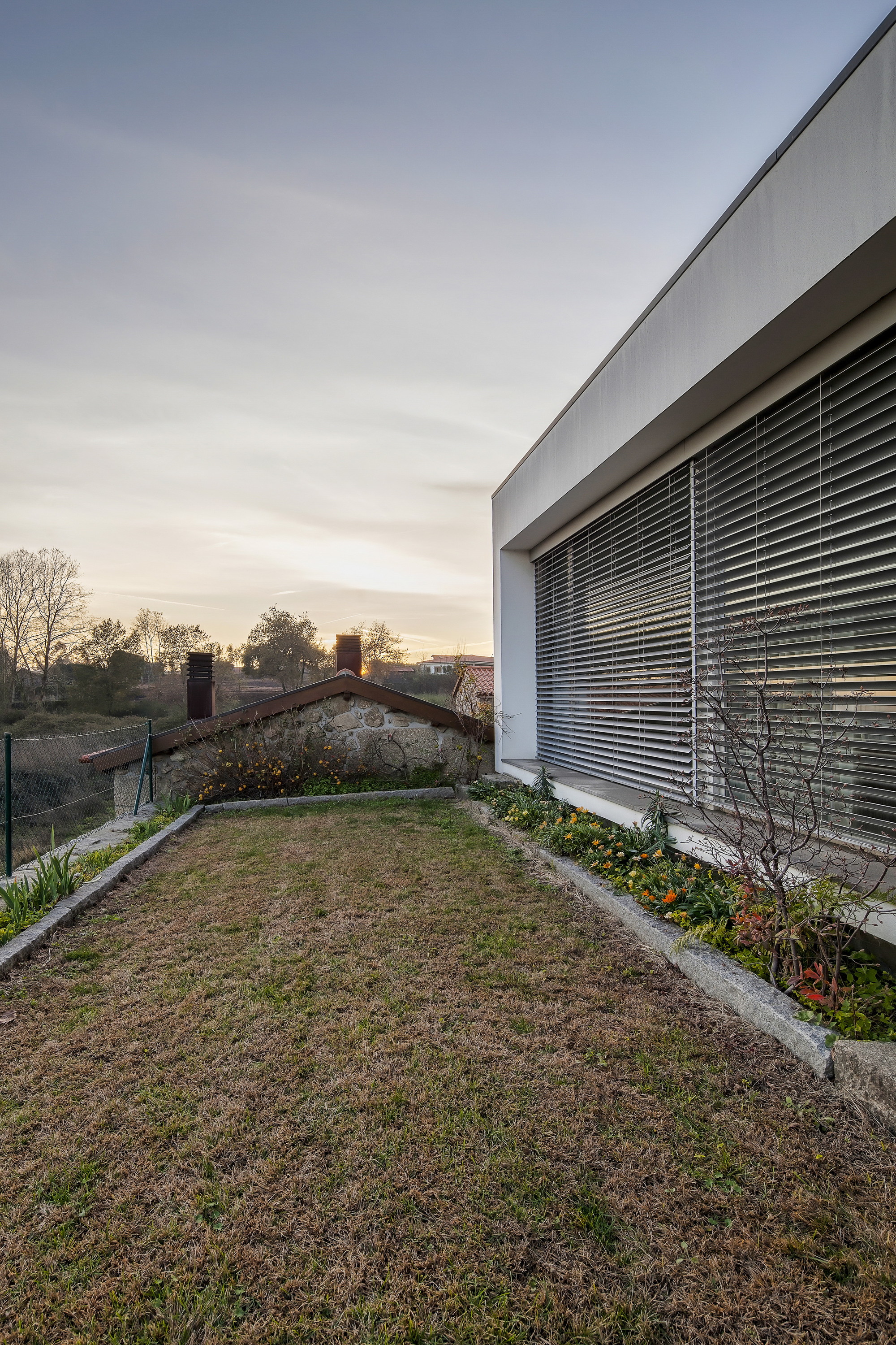 Side of the house with louvred blinds beside a flower bed, a stone outbuilding with chimneys beyond a grass strip at sunset