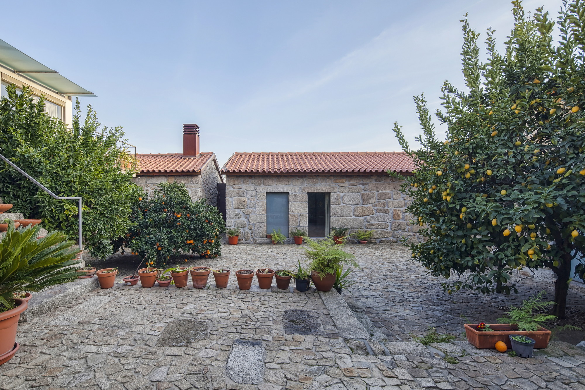 Cobbled courtyard with potted plants and fruiting lemon trees in front of a granite outbuilding with a terracotta tile roof