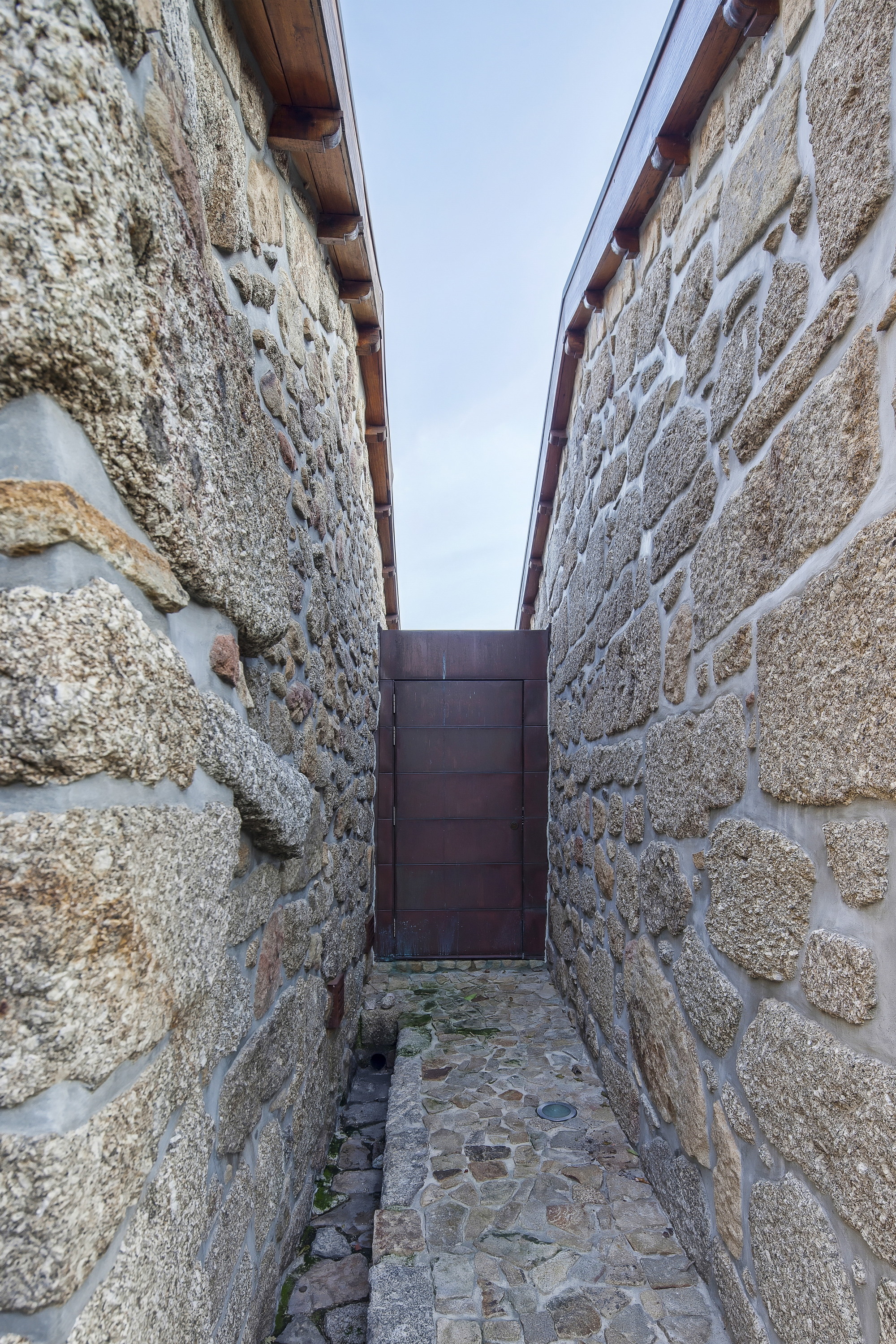 Narrow passage between rough granite walls leading to a rusted steel door, with timber eaves above under a pale sky