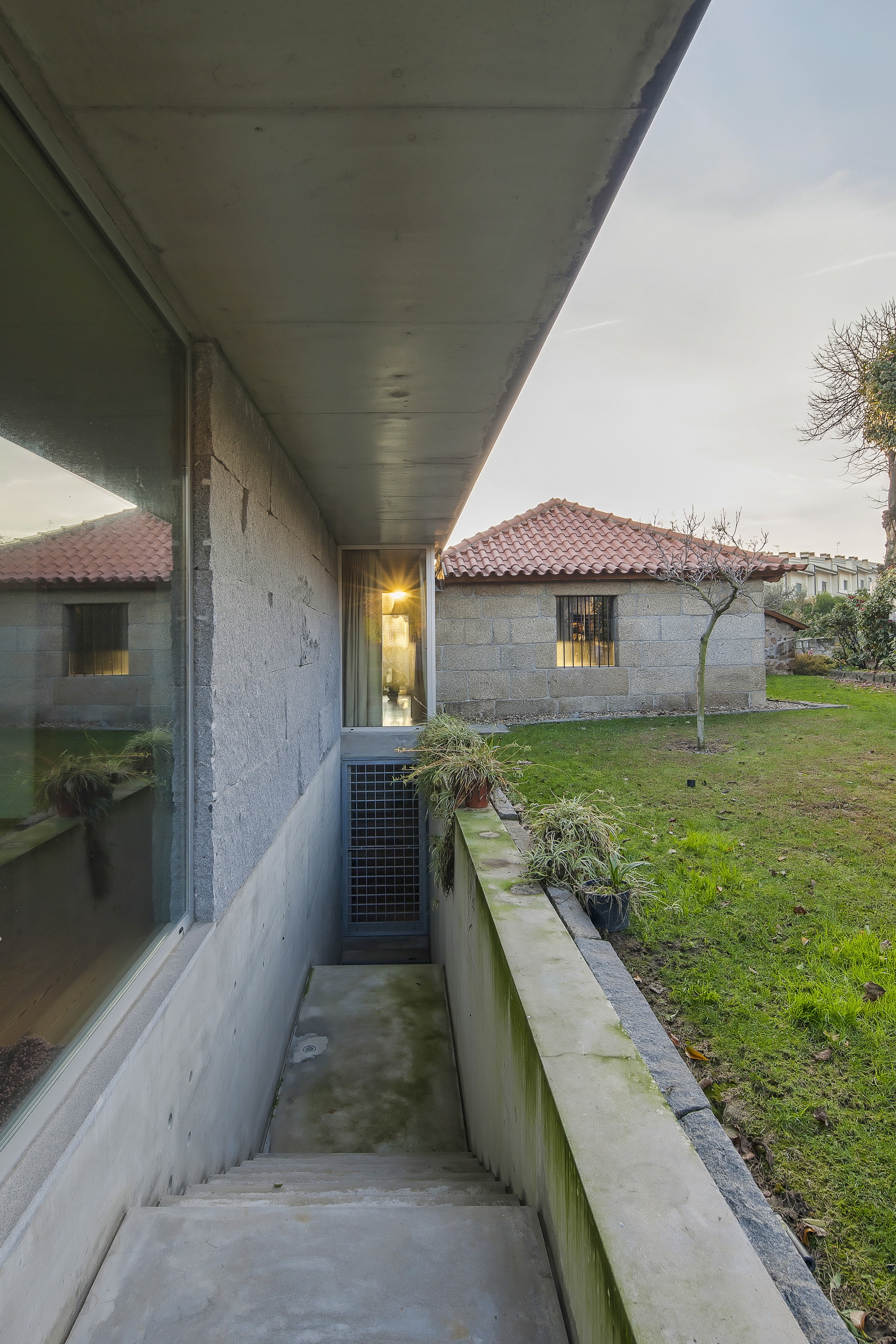 Concrete stair and planted light well beside a glazed wall, with a tiled-roof stone outbuilding across the lawn at dusk