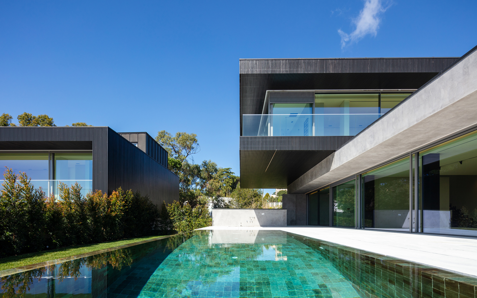 Green glazed tile pool in front of a cantilevered black timber-clad upper volume and concrete canopy under a clear blue sky