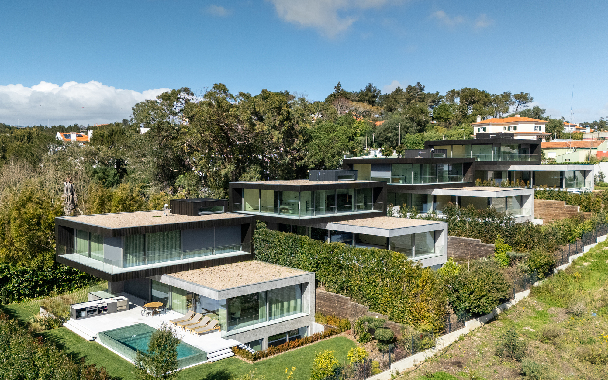 Aerial view of stepped house volumes with flat roofs, glass balconies and a small pool, set among tall trees on a hillside