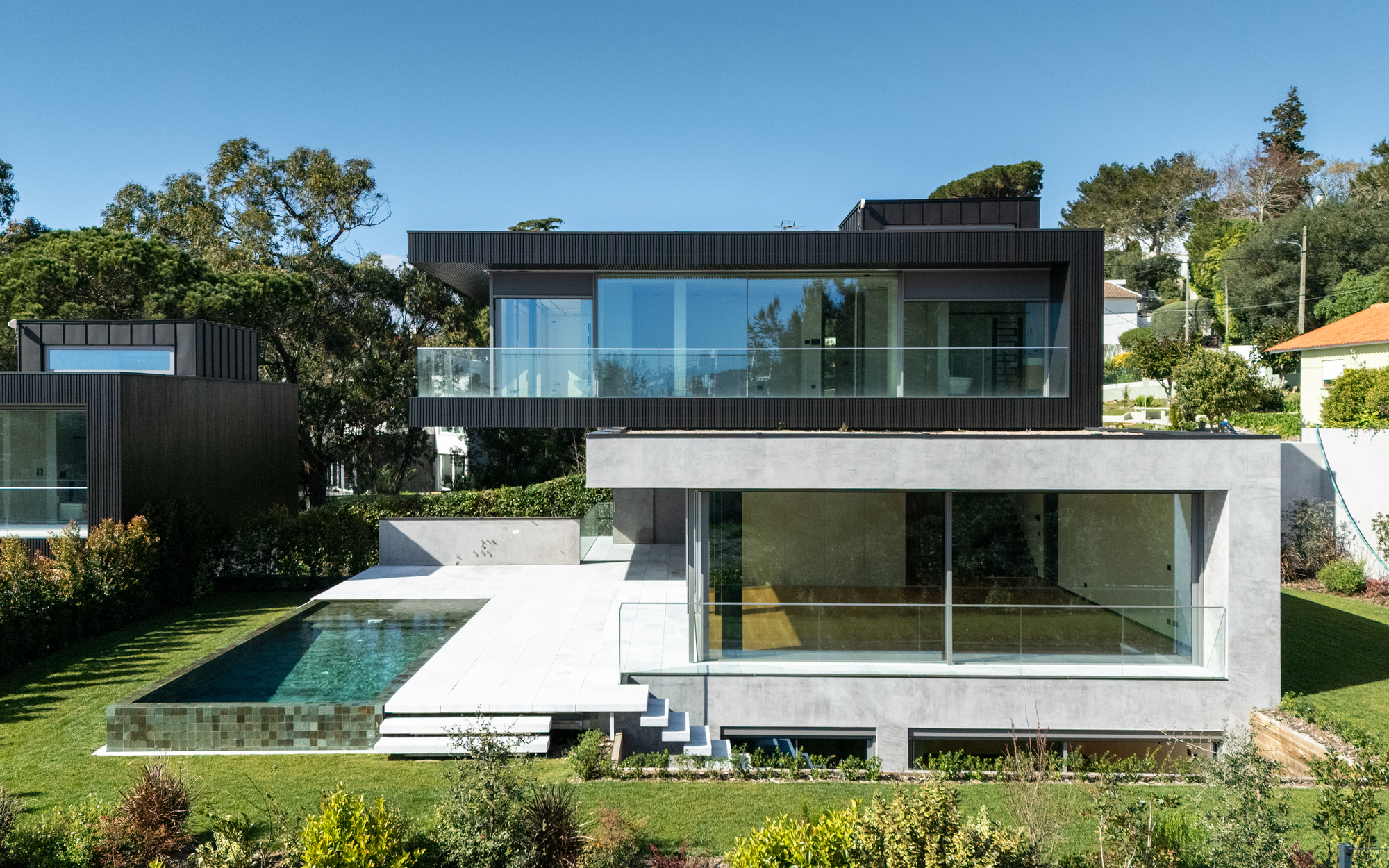 Garden facade with a black clad upper floor over a concrete base, an infinity pool and a pale stone terrace in afternoon sun
