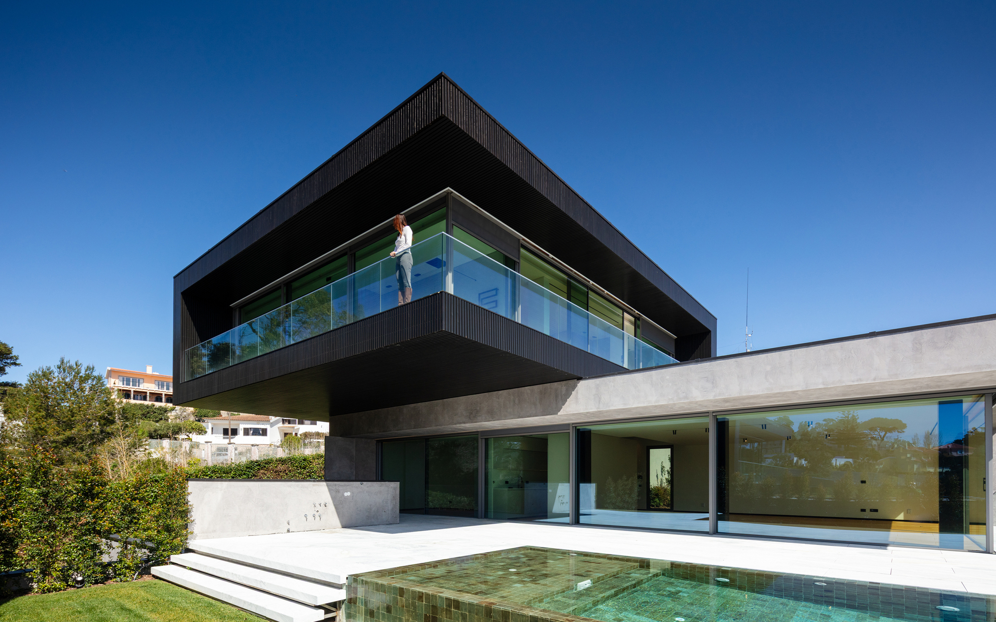 Person standing on a glass-balustraded balcony beneath the dark overhanging box, beside a green tiled pool and sliding glass doors