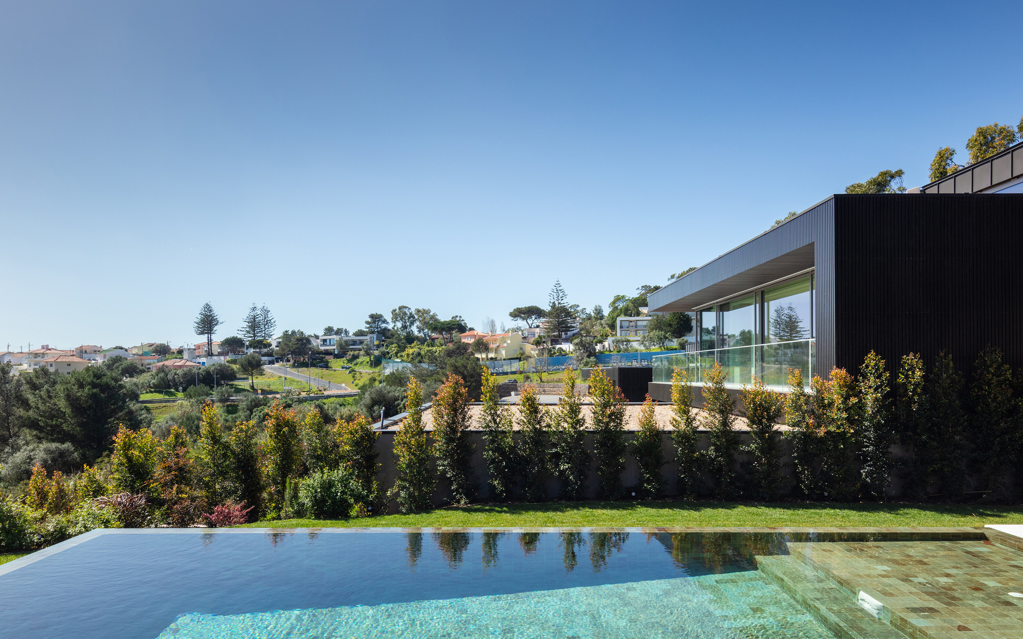 Pool edge with green tile and a row of shrubs, looking past a black clad pavilion toward houses on the distant hillside