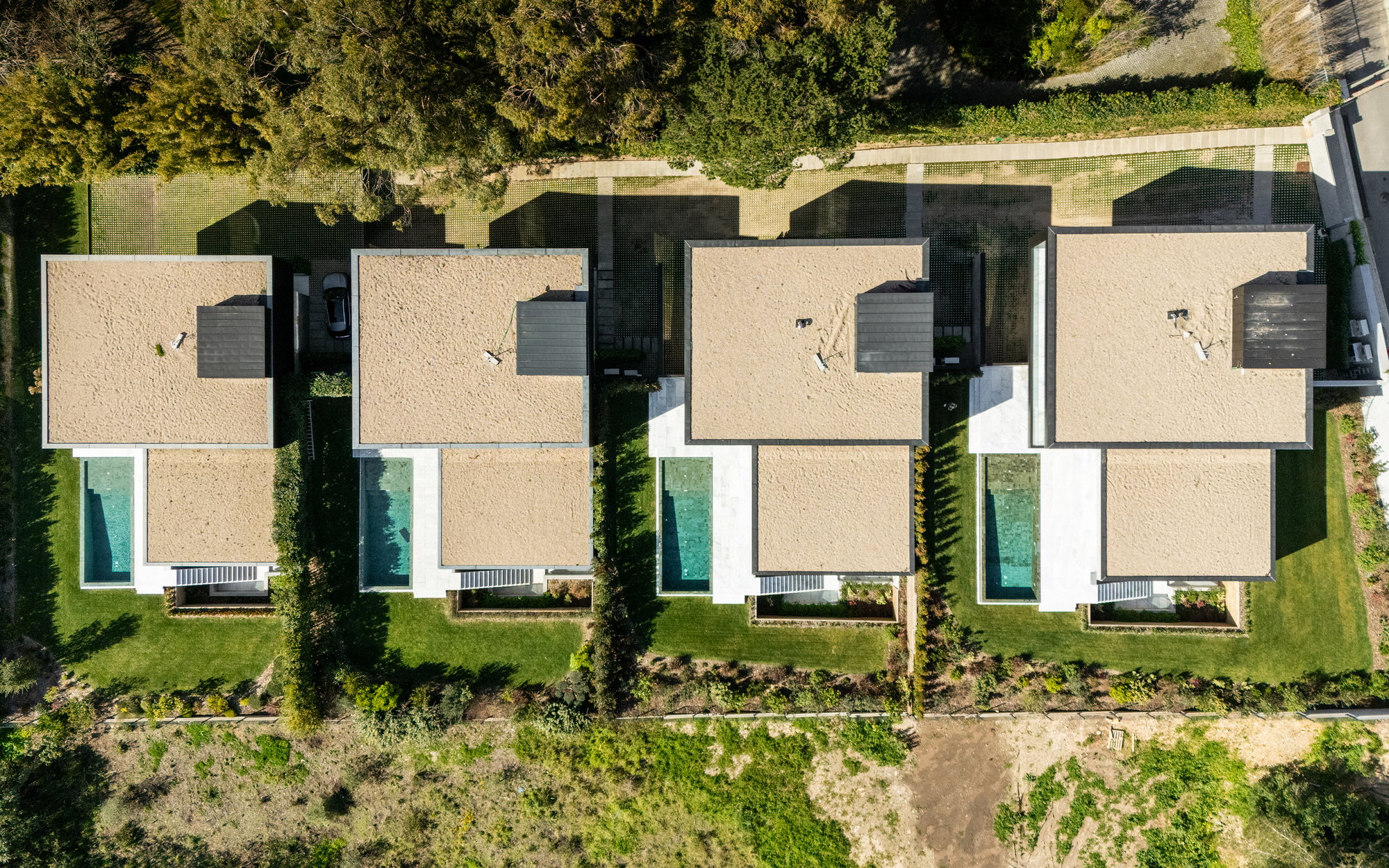 Top-down view of four flat-roofed houses with gravel roofs, green lawns and narrow turquoise pools in afternoon sun