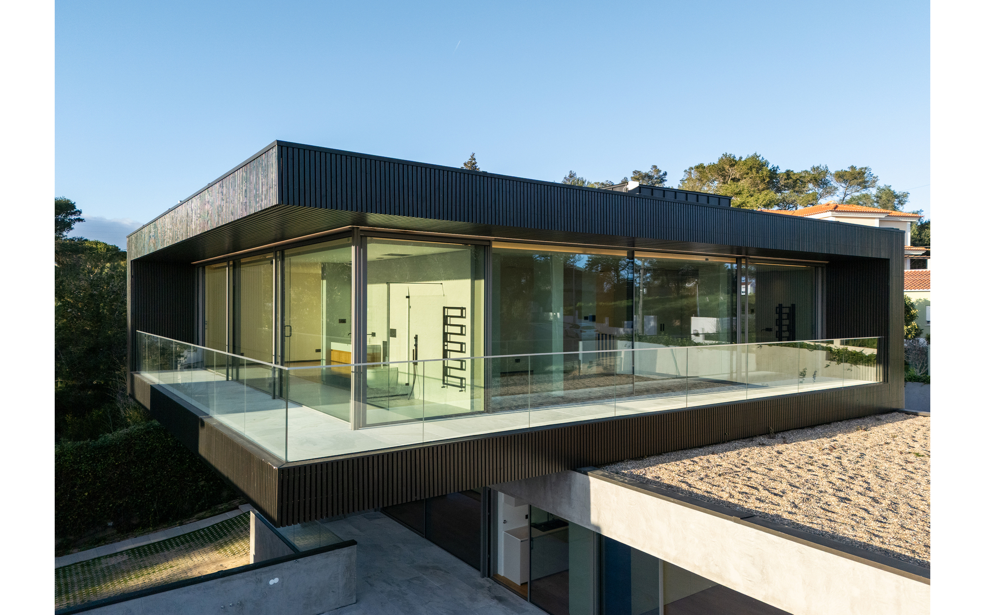 Cantilevered upper volume wrapped in black timber slats with a glass balustrade, beside a gravel roof under clear blue sky