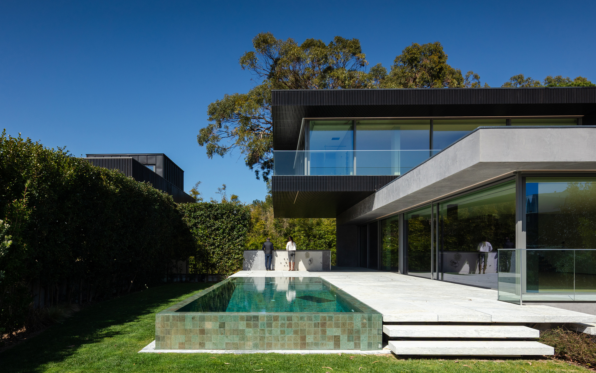 Green-tiled infinity pool beside a stone terrace and steps, with two people standing at a low wall under tall eucalyptus trees